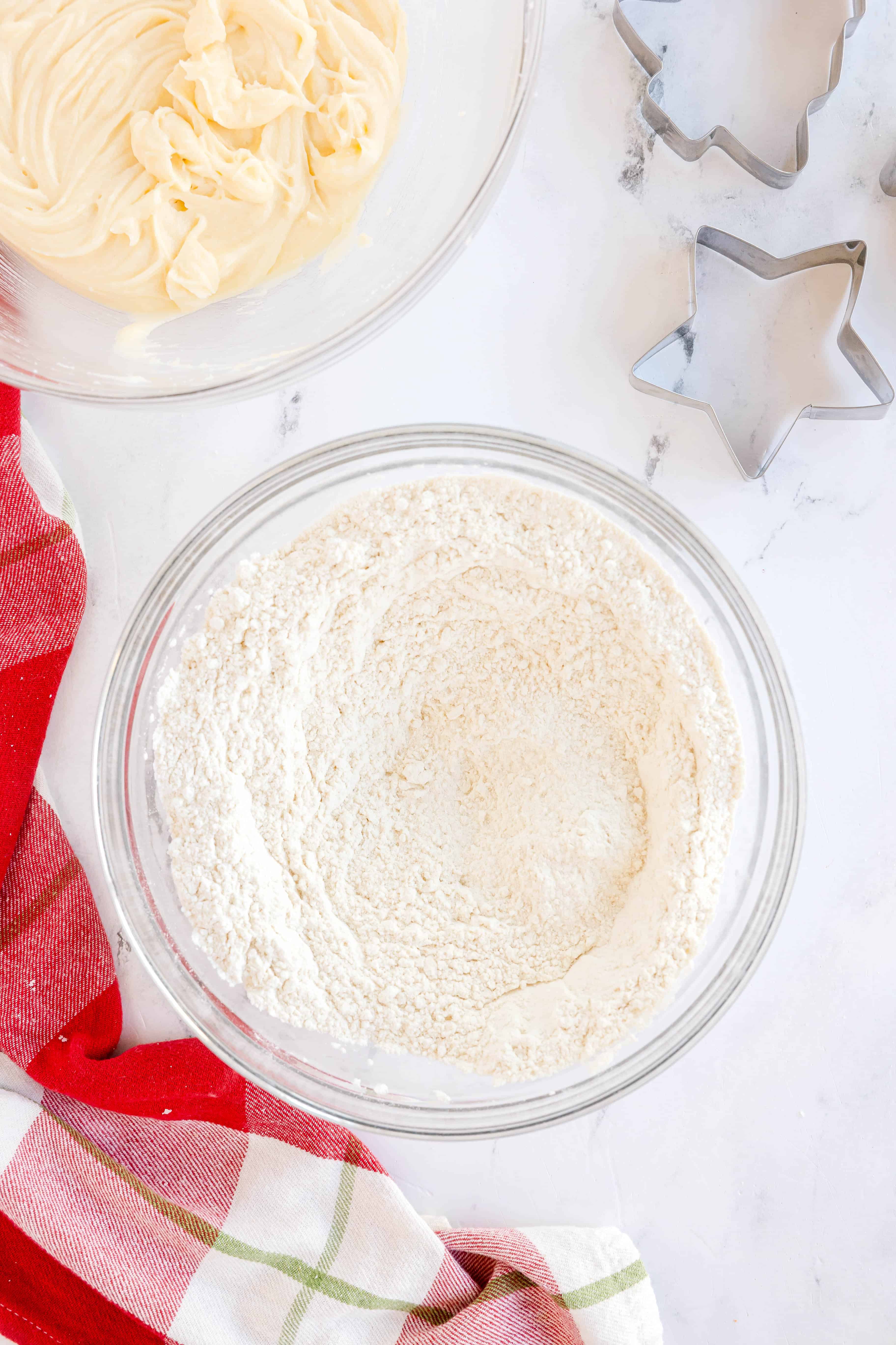 A glass bowl of flour mixture sits on a counter next to a bowl of creamed ingredients, metal cookie cutters, and a red and white plaid cloth.