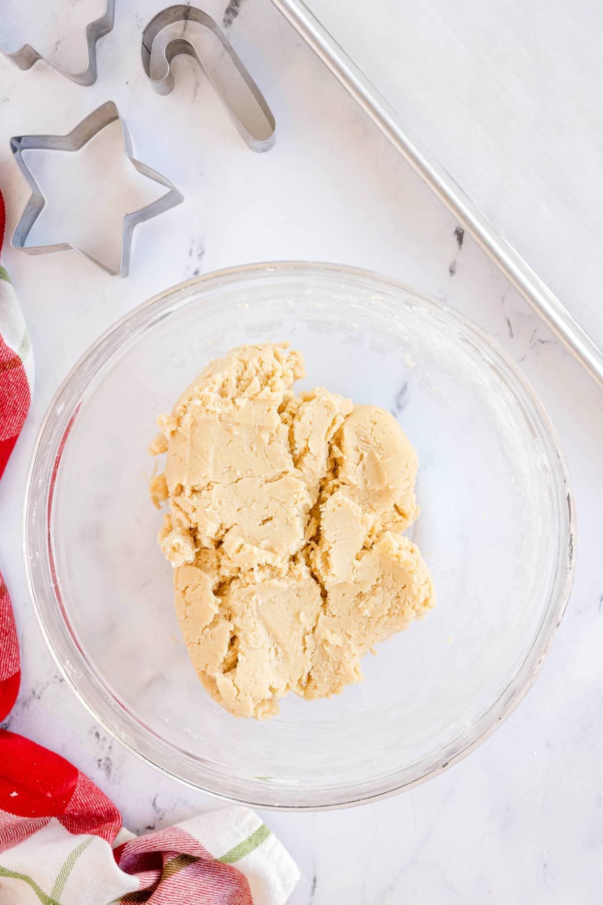A ball of Christmas Sugar Cookies dough in a glass mixing bowl sits on a marble countertop near cookie cutters, a baking sheet, and a red-striped towel.