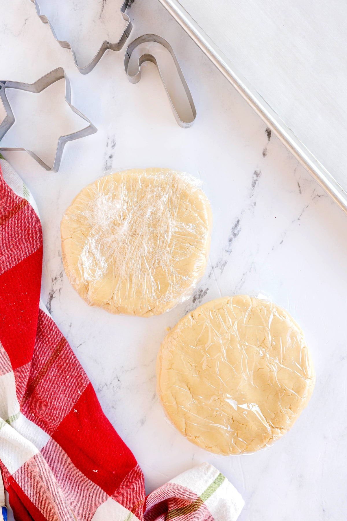 Two discs of Christmas Sugar Cookies dough wrapped in plastic wrap on a marble surface, next to metal cookie cutters, a baking sheet, and a red plaid towel.