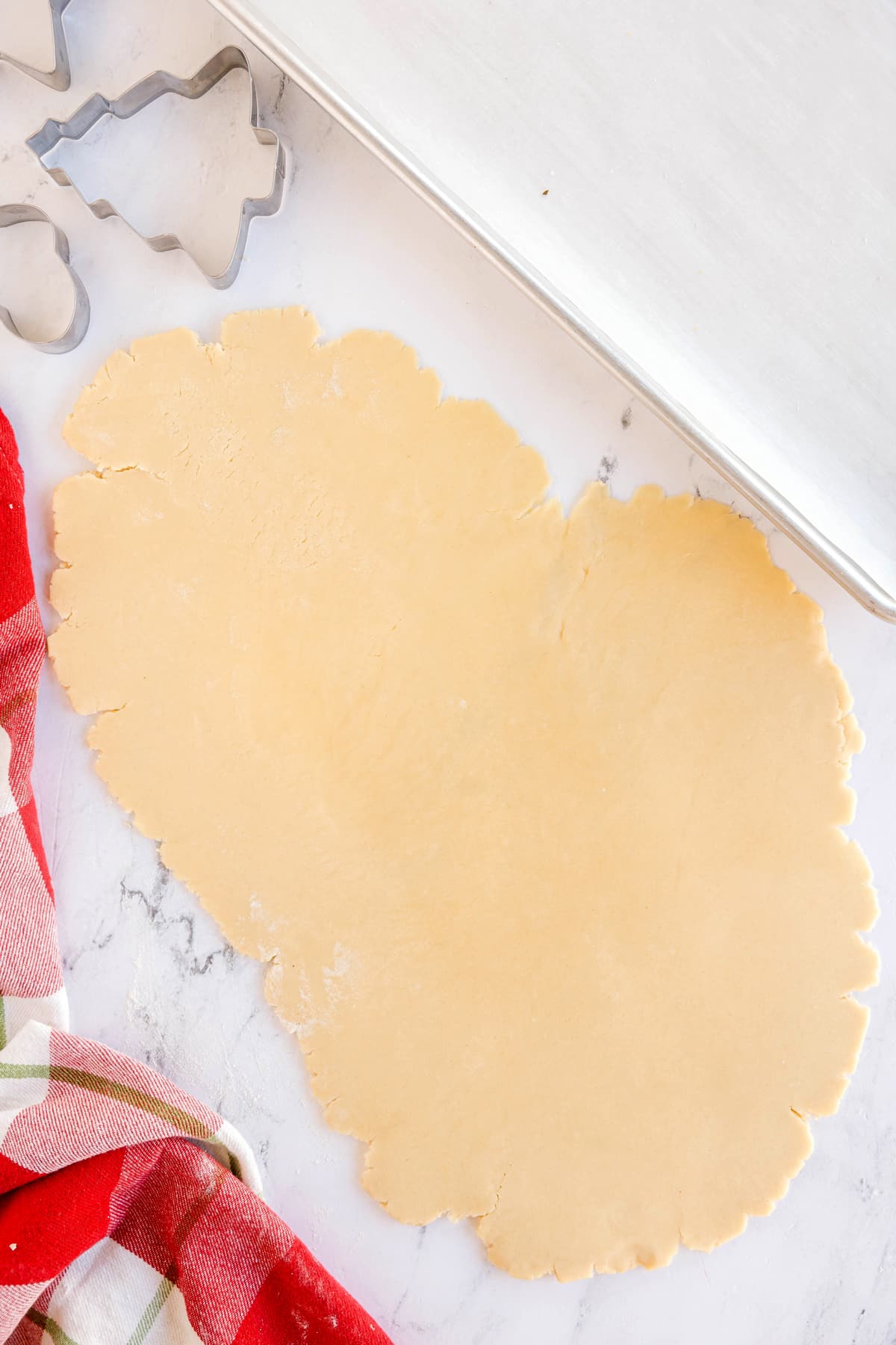 Rolled out Christmas Sugar Cookie dough on a marble surface next to metal cookie cutters, a baking sheet, and a red plaid cloth.