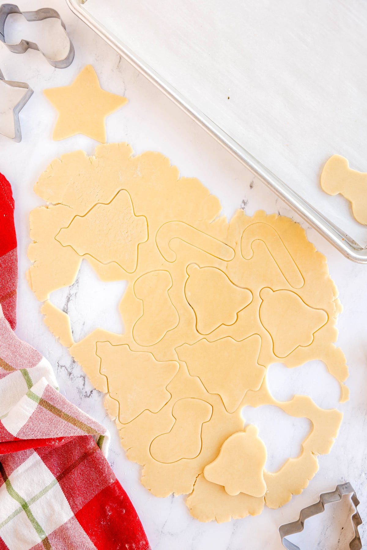 Rolled-out cookie dough with Christmas-themed shapes cut out, surrounded by cookie cutters, a baking sheet, and a red plaid towel on a marble surface. This is one of the many steps in preparing Christmas Sugar Cookies