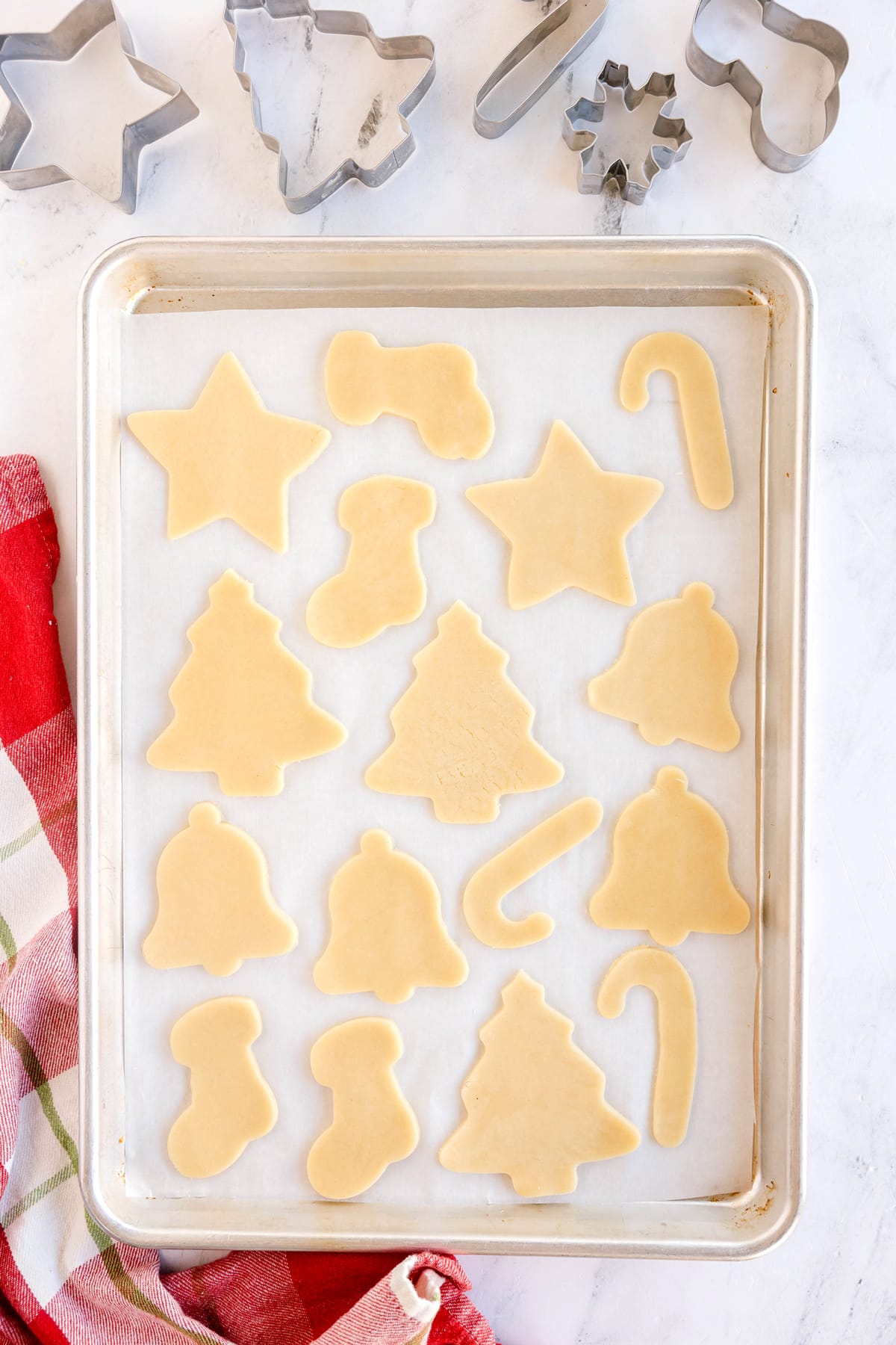 A baking sheet with unbaked Christmas Sugar Cookies in shapes like stars, trees, bells, stockings, and candy canes, alongside cookie cutters and a red plaid towel.