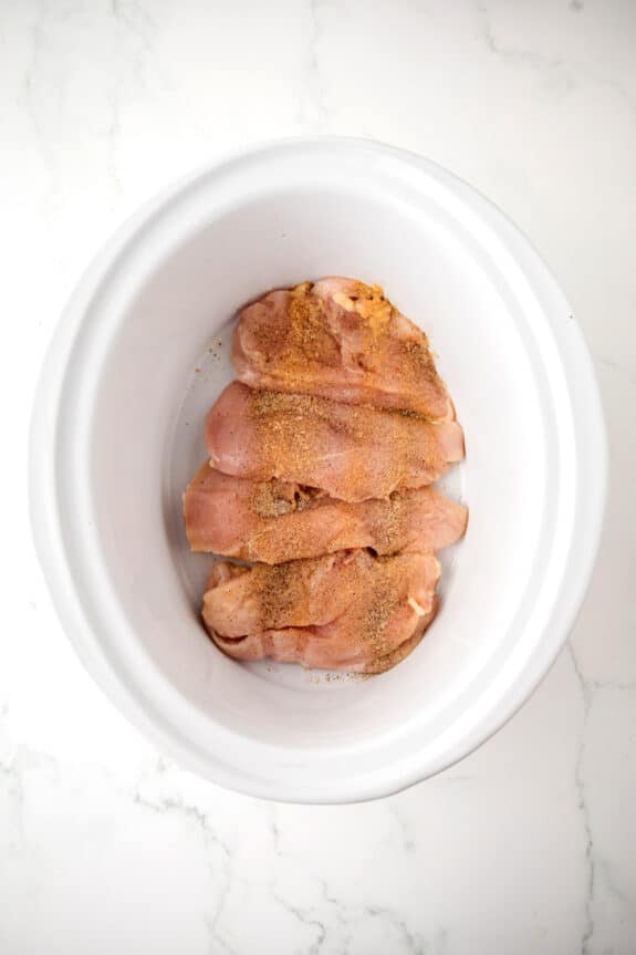 Raw, seasoned chicken breasts are placed in a white oval slow cooker on a white marble surface for Chicken and Stuffing Casserole recipe.