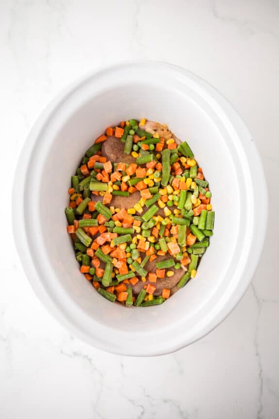A white slow cooker containing Chicken and Stuffing Casserole and a mix of frozen vegetables, including carrots, green beans, and corn, on a white marble surface.