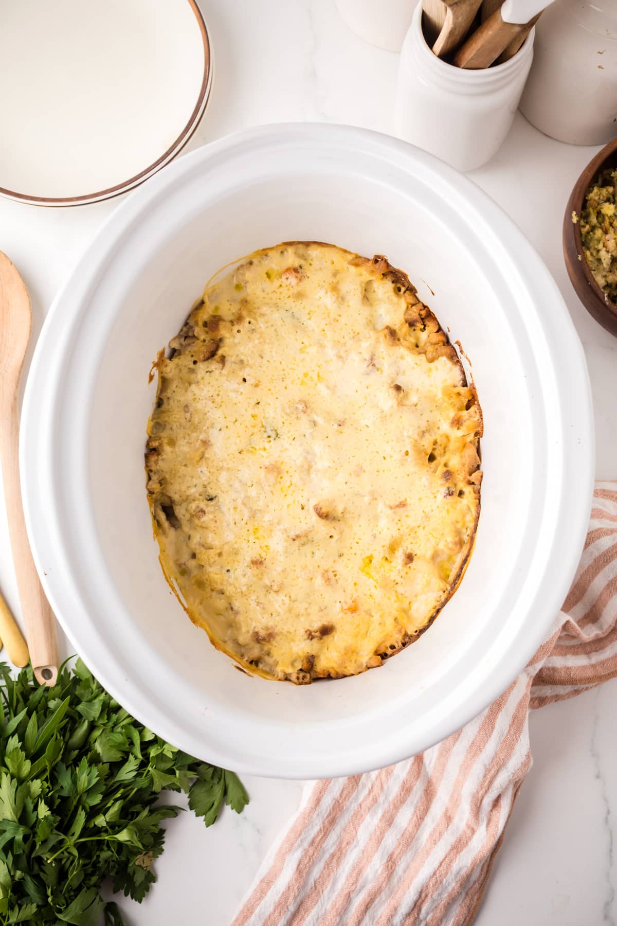 A Chicken and Stuffing Casserole with a browned cheese topping sits in a white oval baking dish on a marble countertop, surrounded by plates, utensils, a striped towel, and fresh parsley.