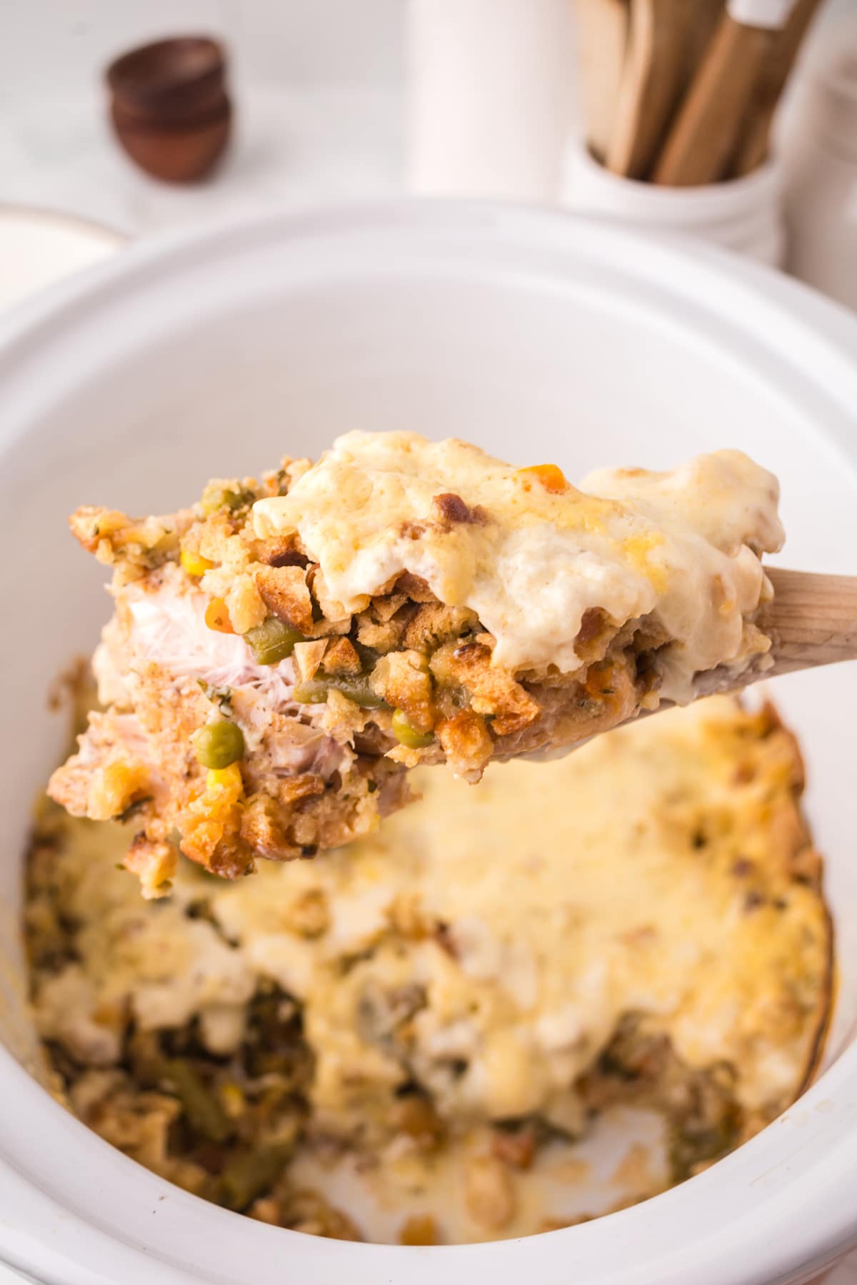 A wooden spoon holds a serving of Chicken and Stuffing Casserole with vegetables and meat above a white baking dish filled with more casserole.