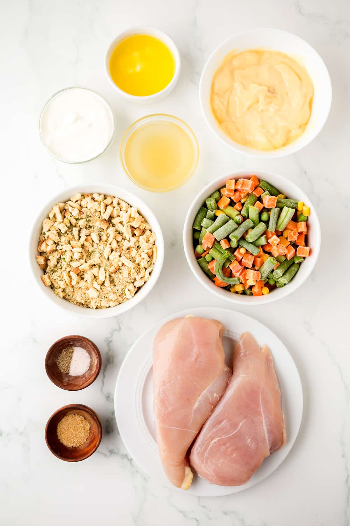 Ingredients for Chicken and Stuffing Casserole are the following: boneless chicken breasts, salt, pepper, poultry seasoning, mixed frozen vegetables, stuffing mix, sour cream, chicken broth, unsalted butter, arranged in separate bowls on a white surface.