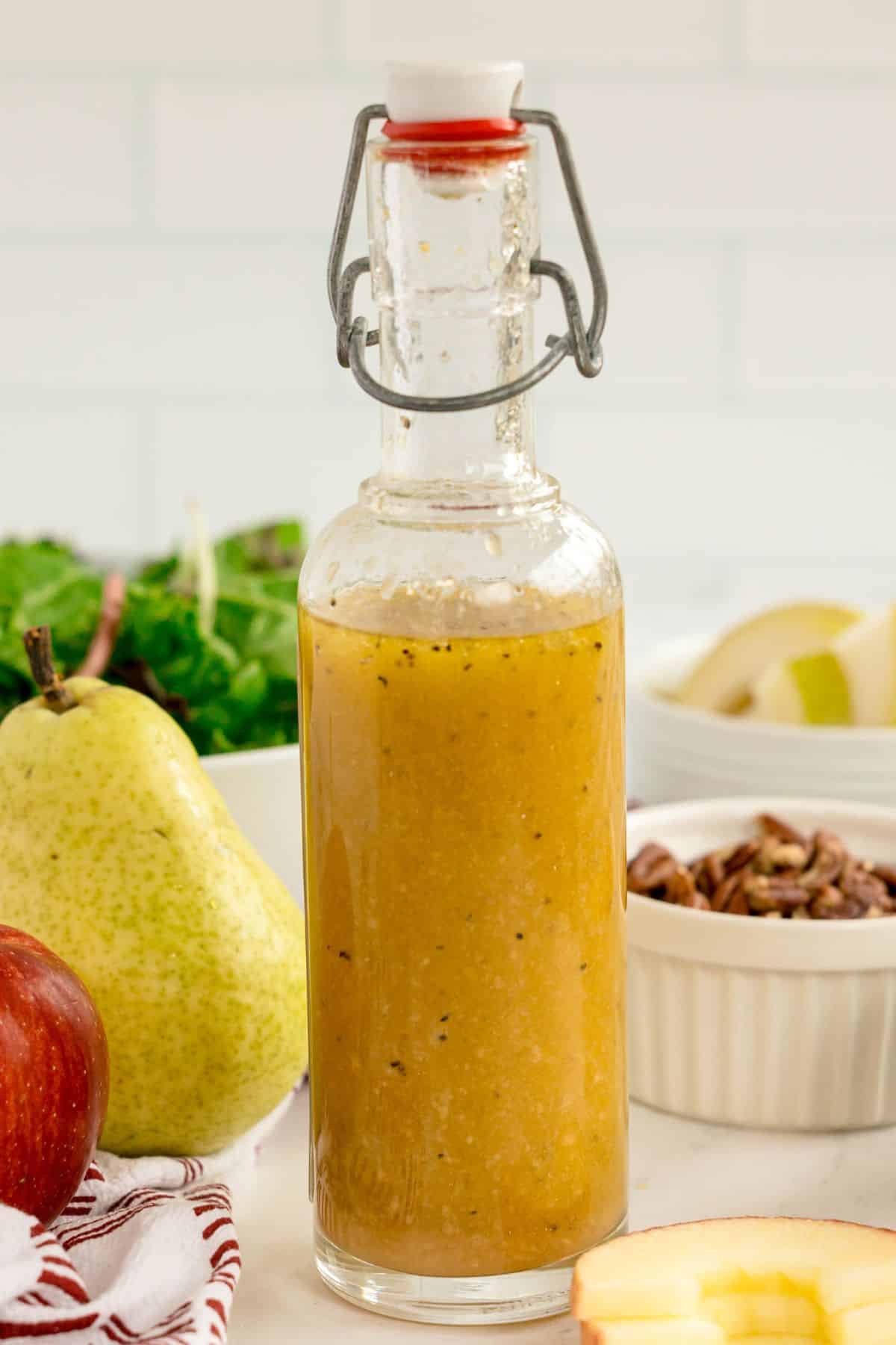 A glass bottle filled with a light brown vinaigrette sits on a counter next to a pear, an apple, salad greens, and chopped pecans—perfect ingredients for a festive Christmas wreath salad.