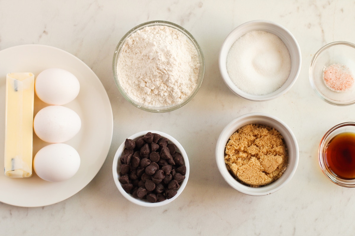 Assorted Fudgy Brownies baking ingredients on a countertop, including unsalted butter, chocolate chips, brown sugar, large eggs, strong coffee, all-purpose flour, and sea salt on small containers