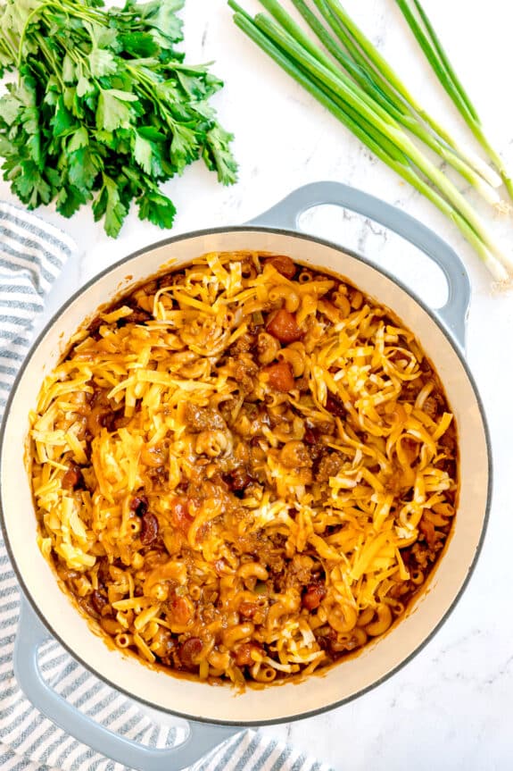A pot of Chili Mac and Cheese with ground beef, beans, and shredded cheese on a marble surface, next to green onions, parsley, and a striped cloth.