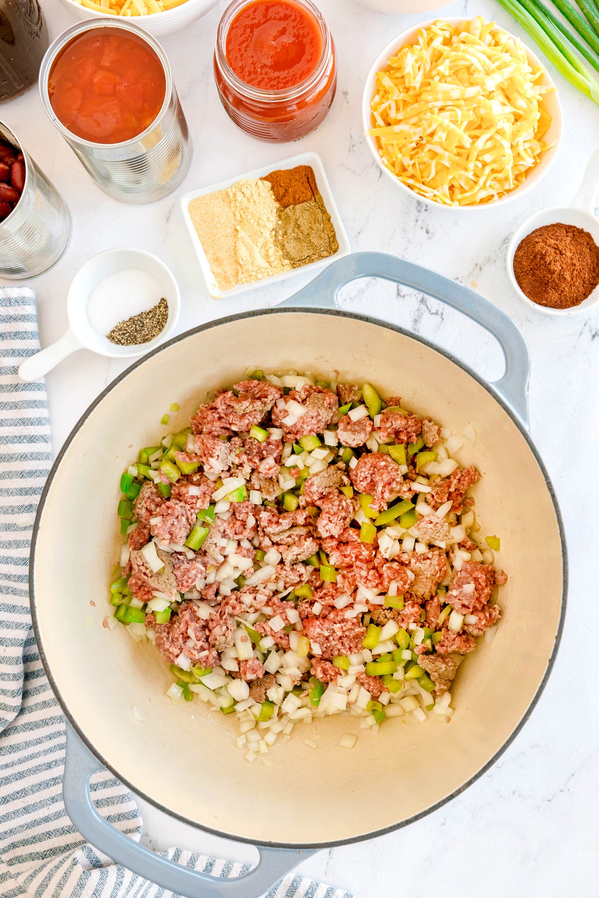 Browning ground meat, chopped onions, and celery in a large pot is the first step in preparing Chili Mac and Cheese. It is surrounded by bowls of spices, canned tomatoes, cheese, and other chili ingredients on a white countertop.