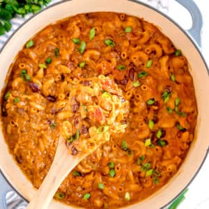 A pot of creamy Chili Mac and Cheese mixed with beans, tomatoes, and garnished with chopped green onions, with a wooden spoon lifting a serving.