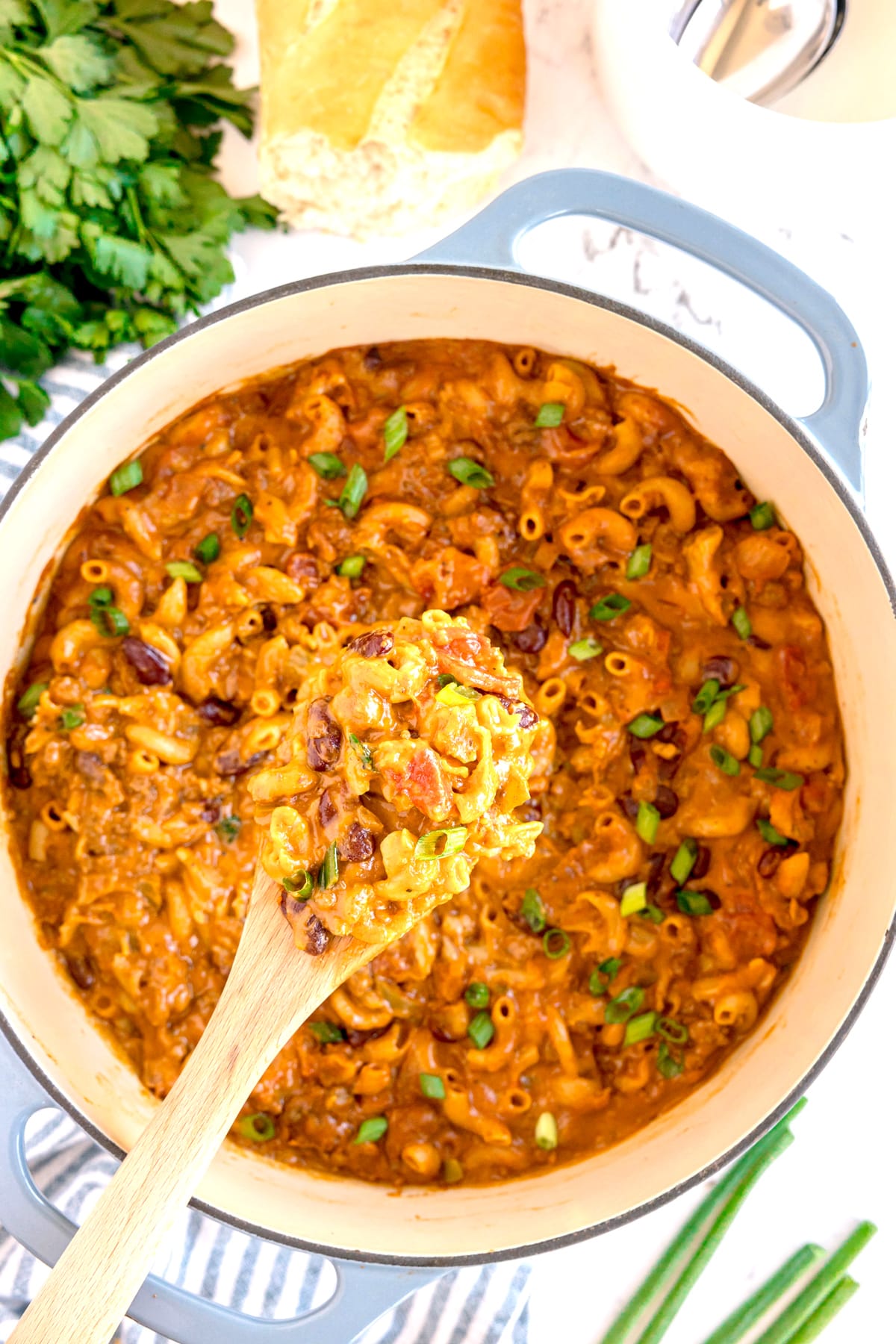 A pot of creamy Chilli Cheese Mac with beans, tomato, and green onions is being stirred with a wooden spoon. Fresh herbs and bread are visible nearby.