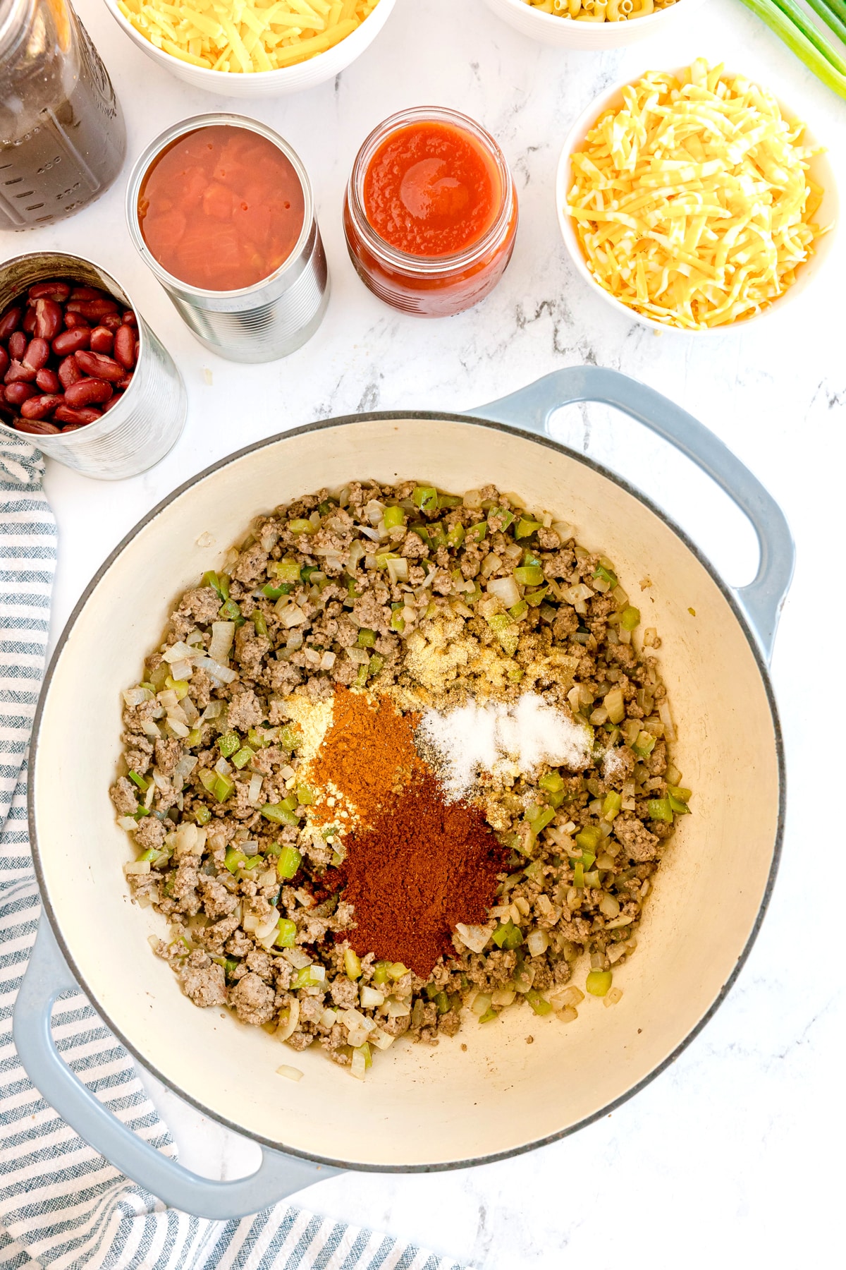 Stir in the garlic powder, chili powder, mustard powder, cumin, smoked paprika, salt, and pepper is nearby are bowls and cans of cheese, beans, tomatoes, and sauce on a white surface. This is just one of the steps in preparing Chili Mac and Cheese