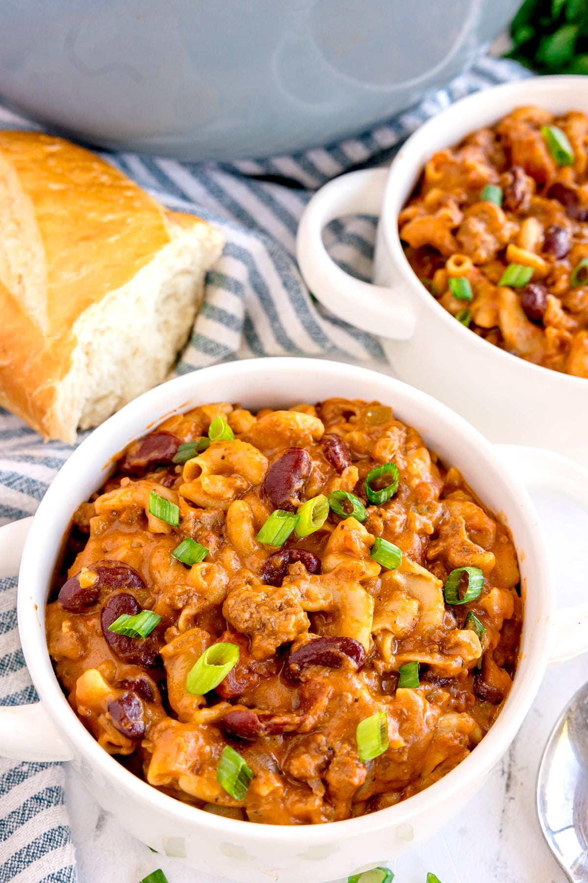 Two bowls of Chili Mac and Cheese topped with chopped green onions, with a sliced baguette and a striped cloth in the background.