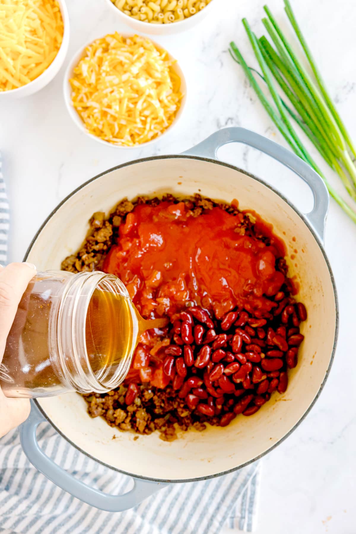 A hand pours beef broth from a jar into a pot containing ground meat, kidney beans, and tomato sauce for Chili Mac and Cheese. Bowls of cheese, pasta, and green onions are nearby on a white surface.