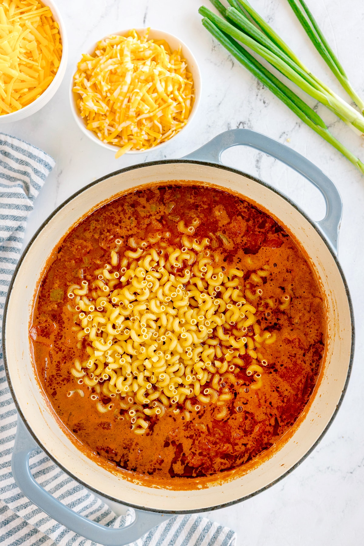 Adding macaroni on pot of tomato mixture for Chili Mac and Cheese, next to bowls of shredded cheese and green onions on a marble countertop.