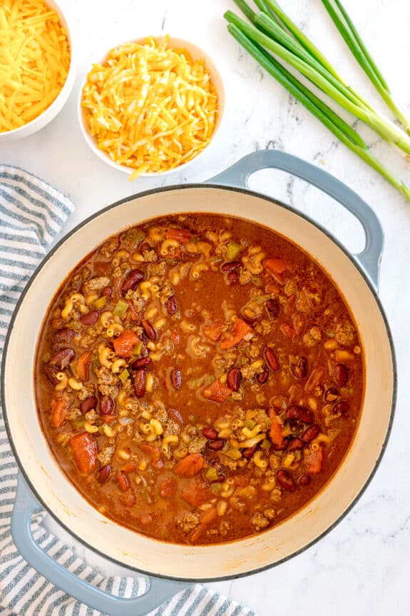 A pot of Chili Mac and Cheese with macaroni, ground meat, kidney beans, and tomatoes next to bowls of shredded cheese and green onions on a marble surface.