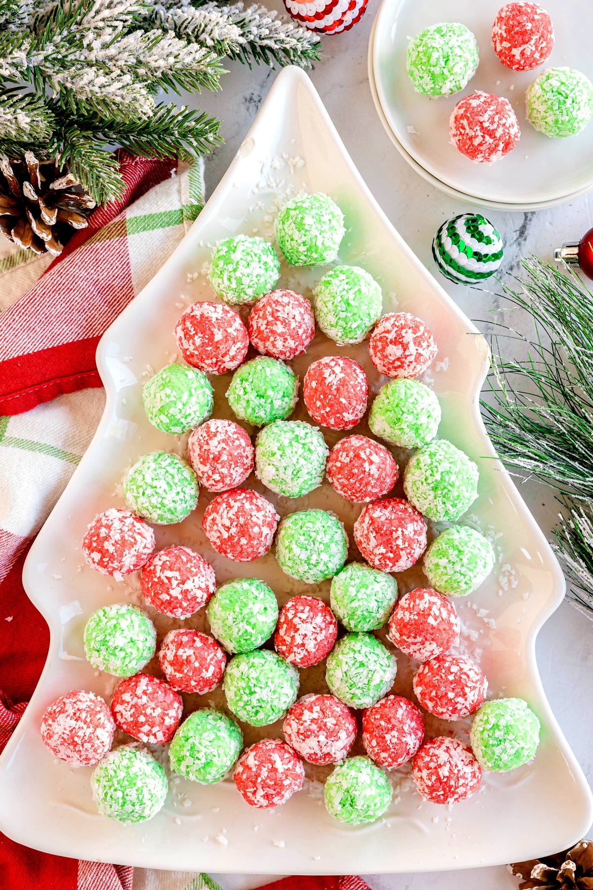A tree-shaped platter holds red and green Christmas Jello Balls arranged in rows, with pine branches and holiday decorations nearby.