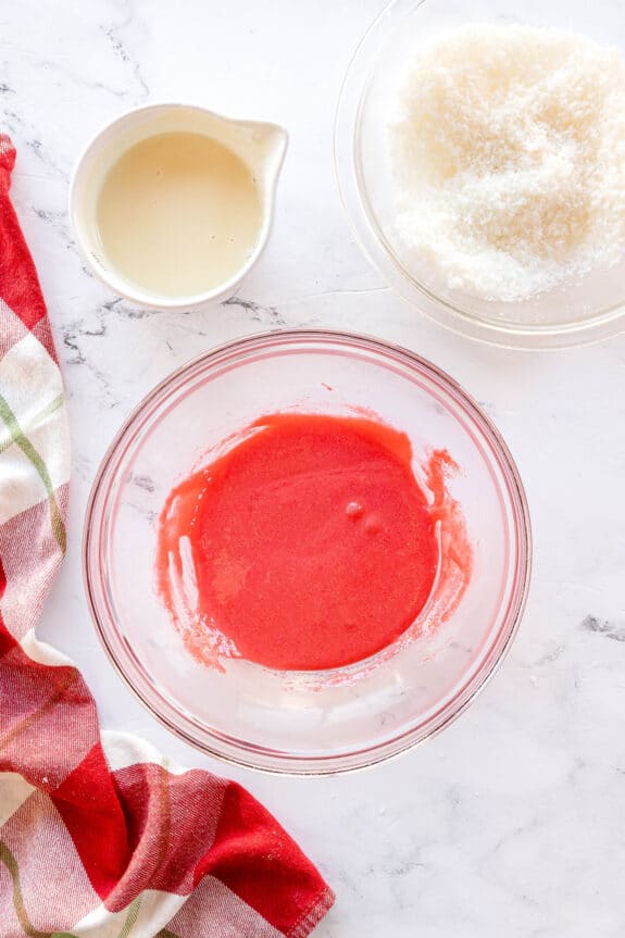 First step in preparing Christmas Jello Balls is to use a clear glass bowl with red liquid, a bowl of shredded coconut, a small pitcher of cream, and a red plaid towel on a white surface.