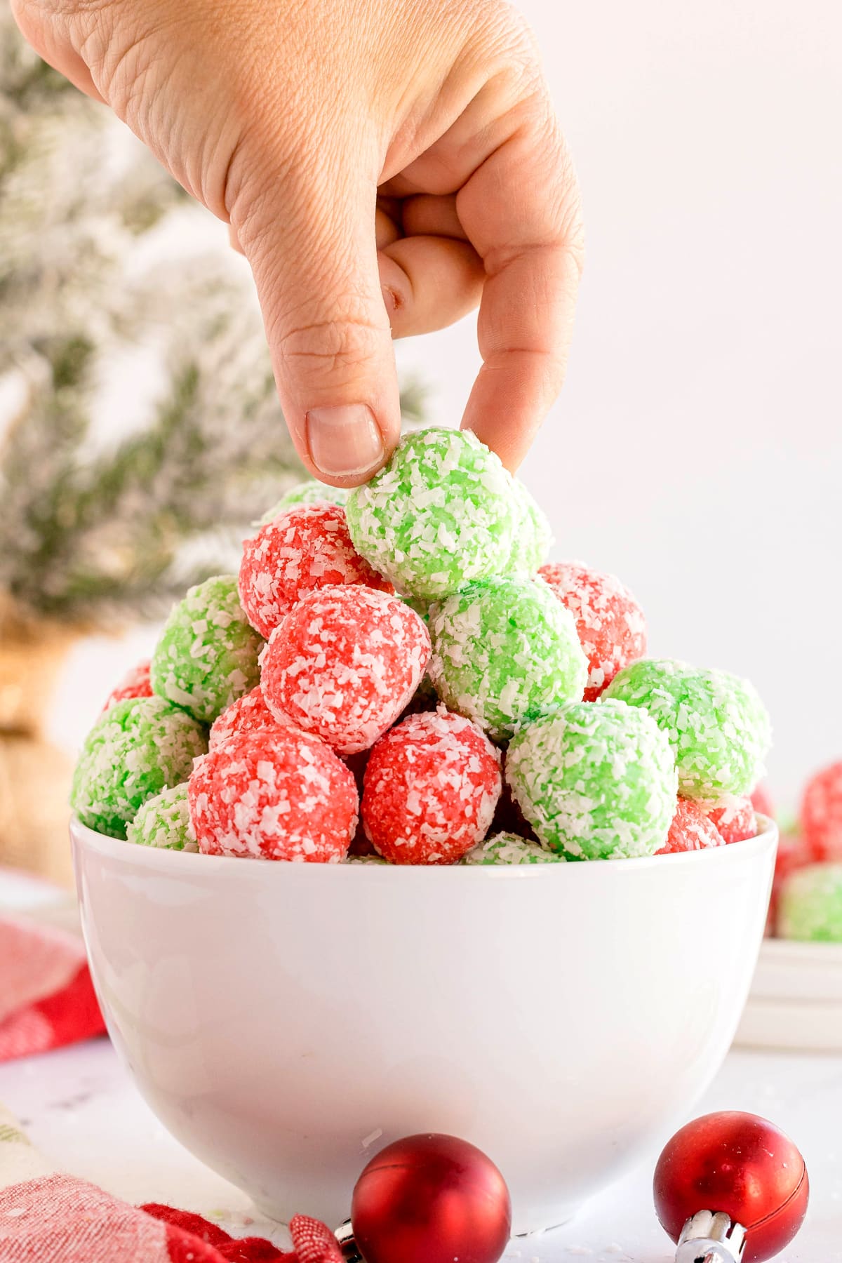 A hand picks up a piece of Christmas Jello Balls from a white bowl filled with similar treats, with festive decor in the background.