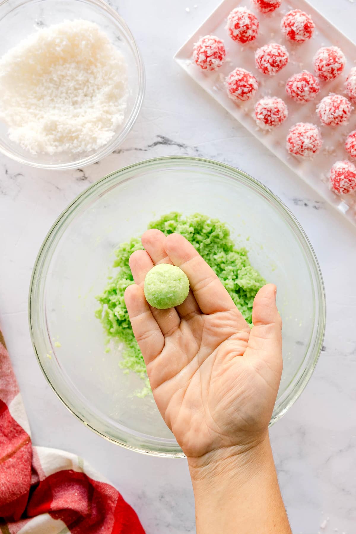 A hand holds a green Christmas Jello Balls over a glass bowl of green mixture, with a bowl of shredded coconut and a tray of red coconut-covered balls nearby.