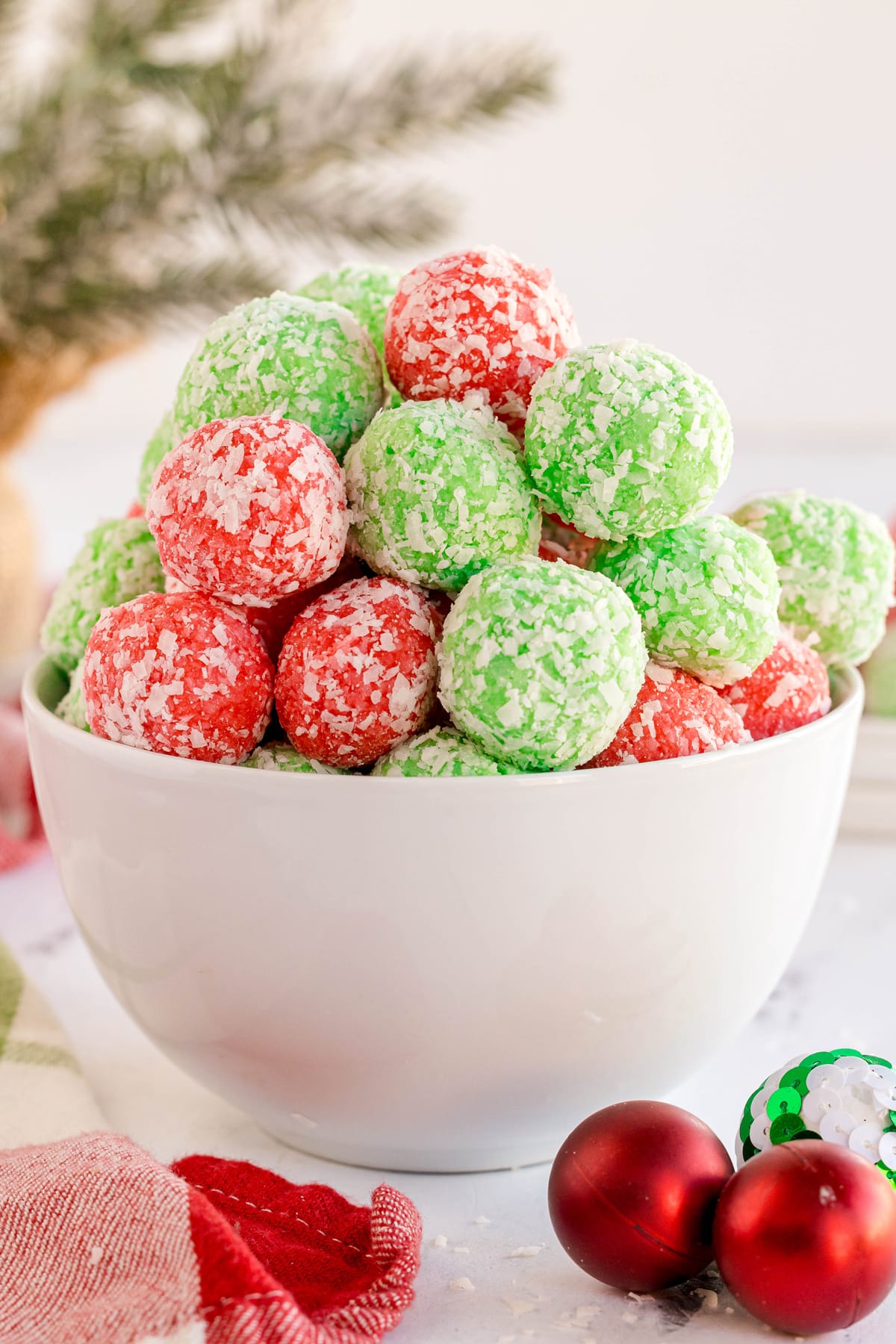 A white bowl filled with red and green coconut-covered balls, with Christmas ornaments and greenery in the background.