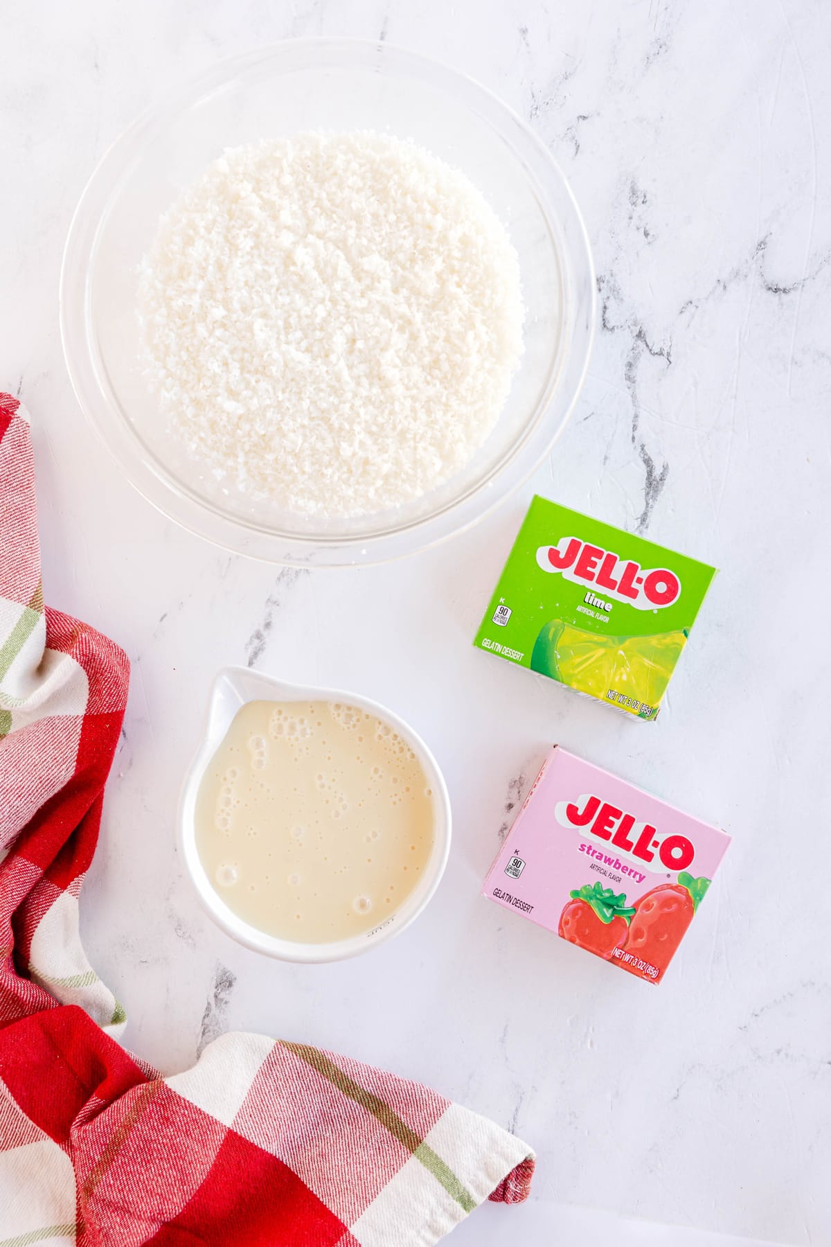 Ingredients of Christmas Jello Balls include a bowl of red jello, green jello, sweetened condensed milk, and finely grated sweetened coconut, on a white surface with a red-checked cloth.