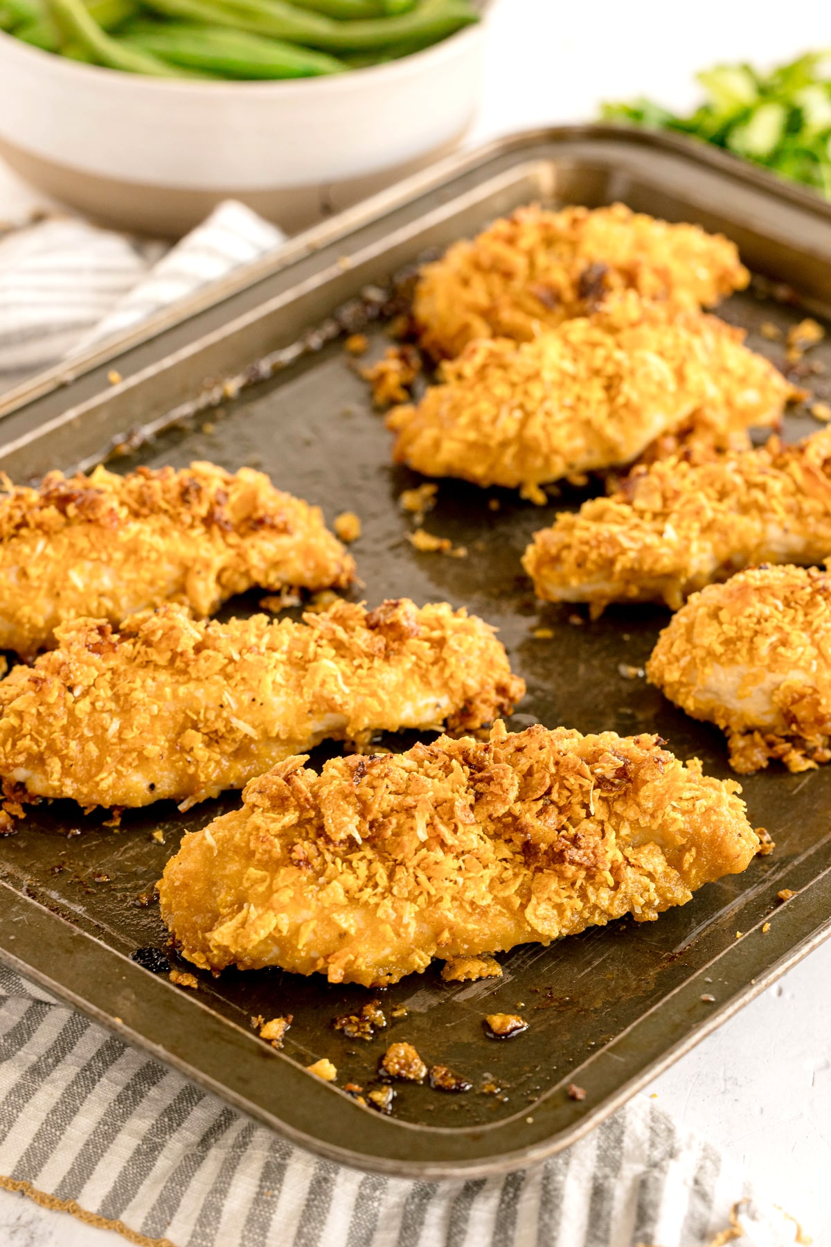A baking tray with several pieces of golden, crispy baked Cornflake Chicken resting on it, with a bowl of green beans blurred in the background.