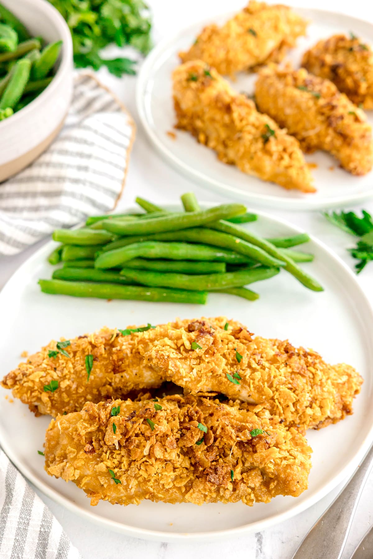 Two plates with crispy Cornflake Chicken tenders and a side of green beans are arranged on a white surface, with a bowl of green beans in the background.