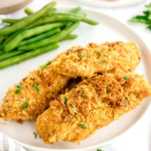 Two crispy Cornflake Chicken tenders on a white plate, garnished with herbs, served with a side of green beans.