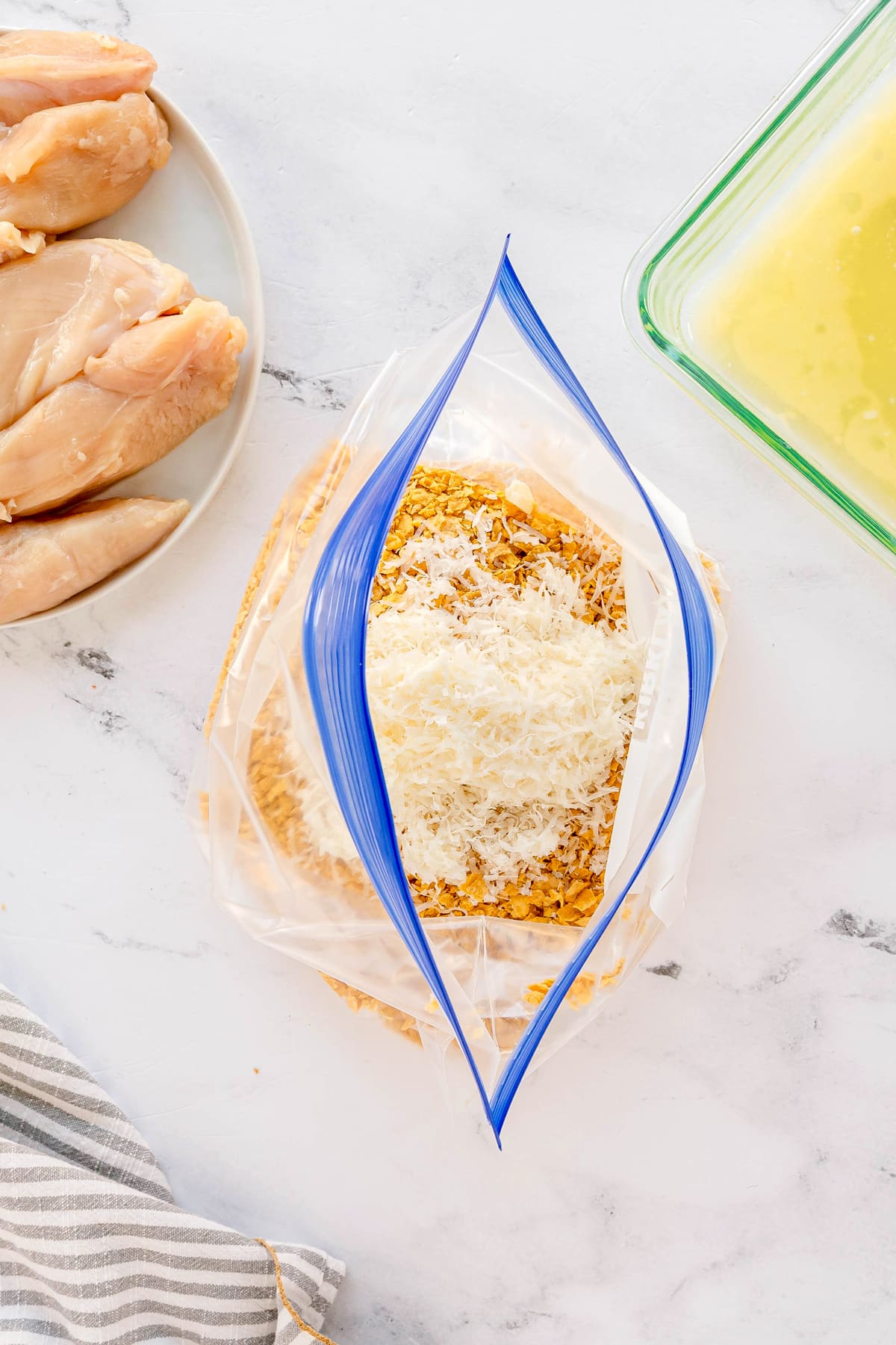 A resealable plastic bag filled with crushed cornflakes, Parmesan cheese, salt, pepper, and garlic powder, sits on a counter near raw chicken breast and a dish of beaten eggs. This is the first step in preparing Cornflake Chicken
