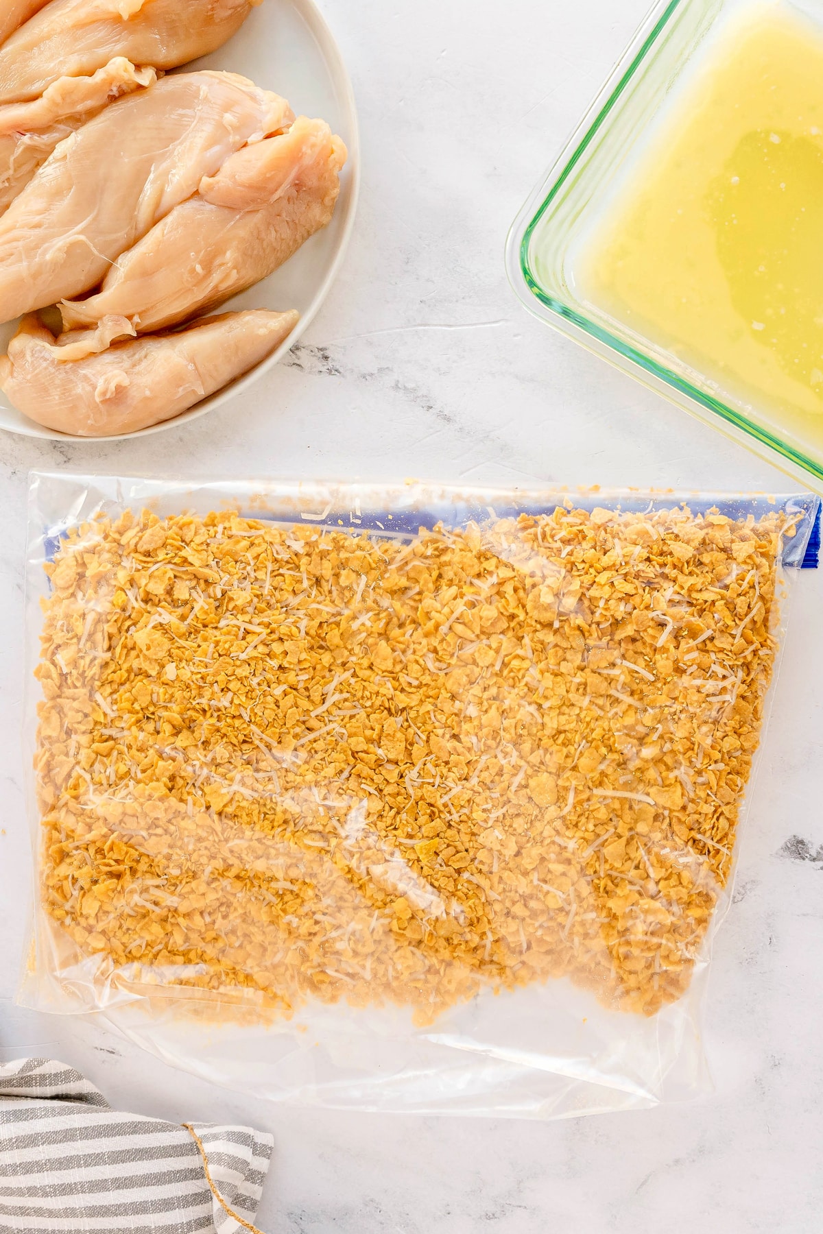 Cornflake Chicken recipe starts in mixing crushed cornflakes and chicken in a sealed plastic bag. A glass dish with beaten eggs on a light countertop can also be seen.