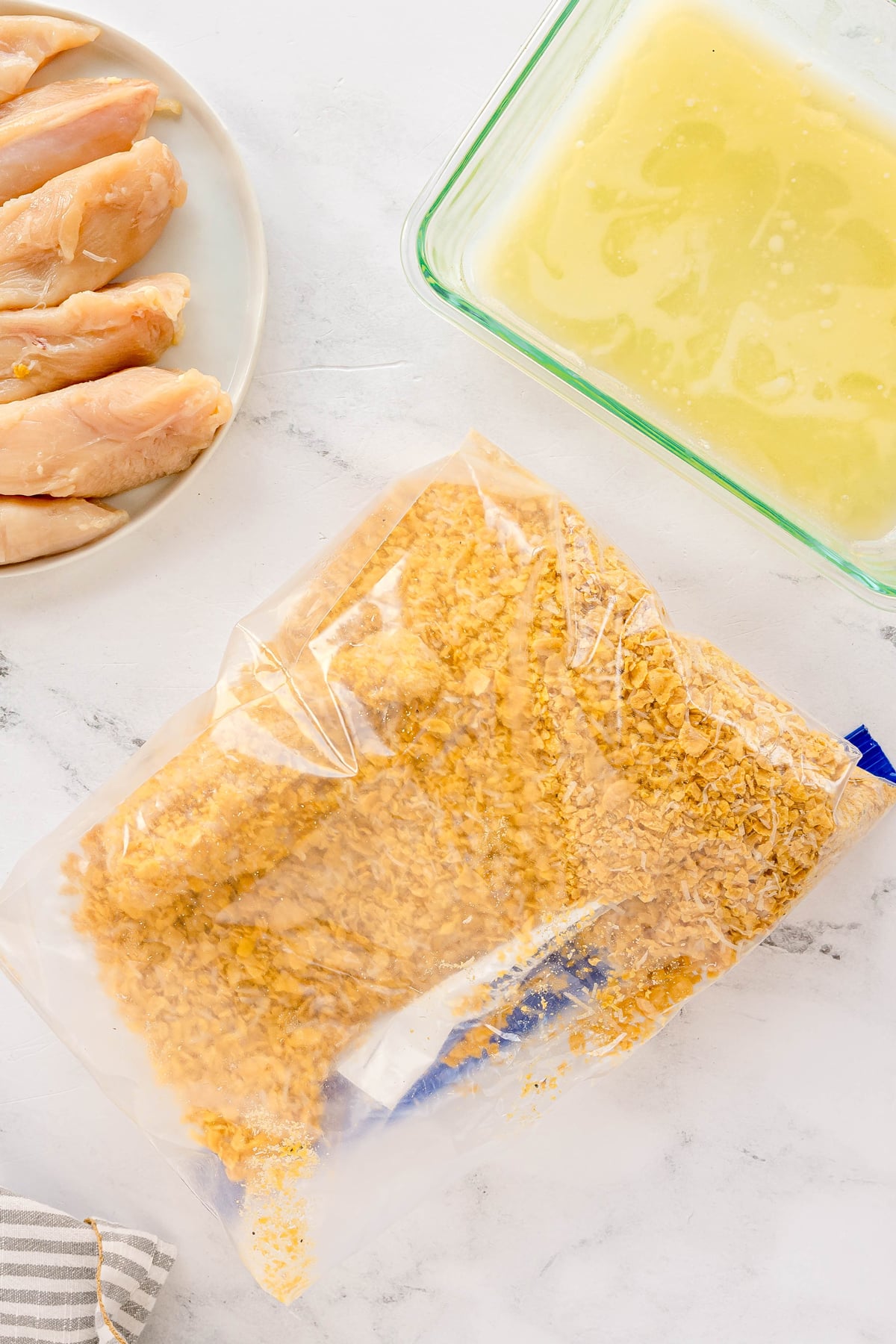 Raw chicken strips on a plate, a dish of liquid, and a plastic bag filled with crushed cornflakes are arranged on a white surface. This is for Cornflake Chicken recipe.