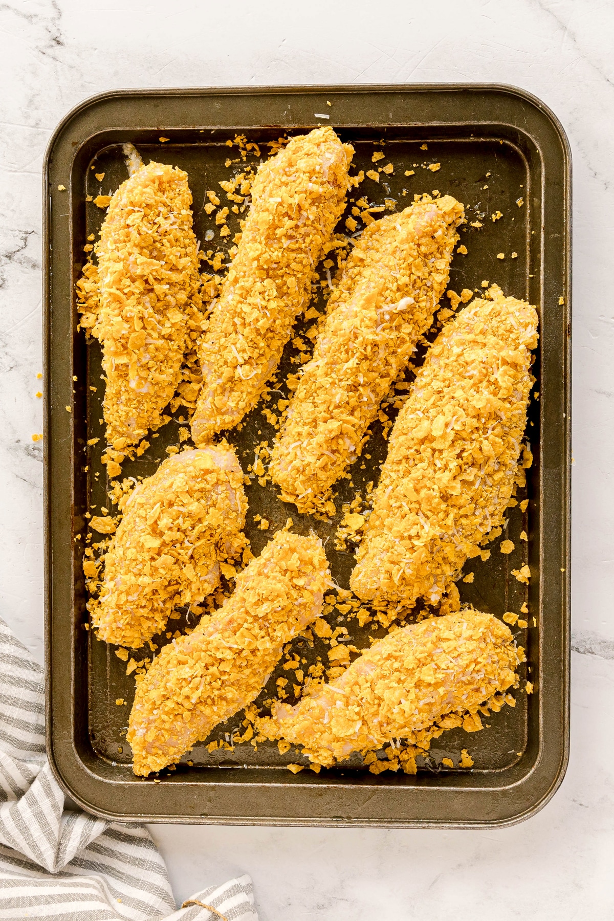 Seven breaded Cornflake Chicken coated in crushed cornflakes are arranged on a baking tray, ready to be cooked.
