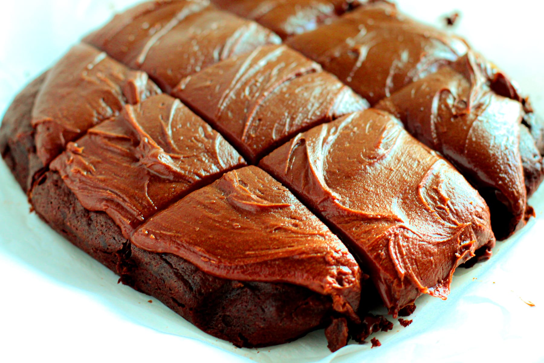 A batch of Fudgy Brownies with a thick layer of chocolate frosting, cut into nine square pieces on parchment paper.