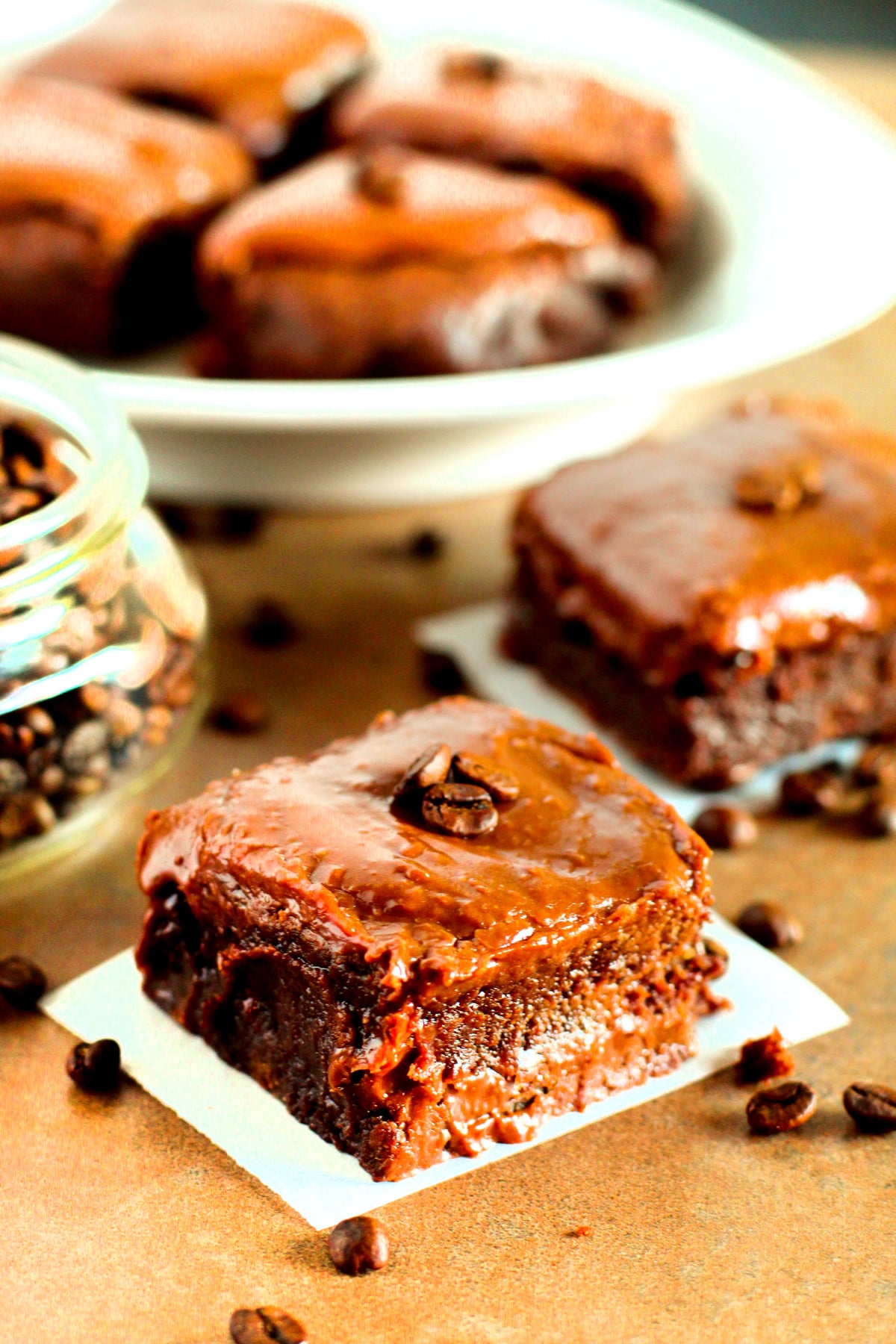 A close-up of Fudgy Brownies with glossy frosting, topped with coffee beans, placed on a table with scattered coffee beans and a jar in the background.