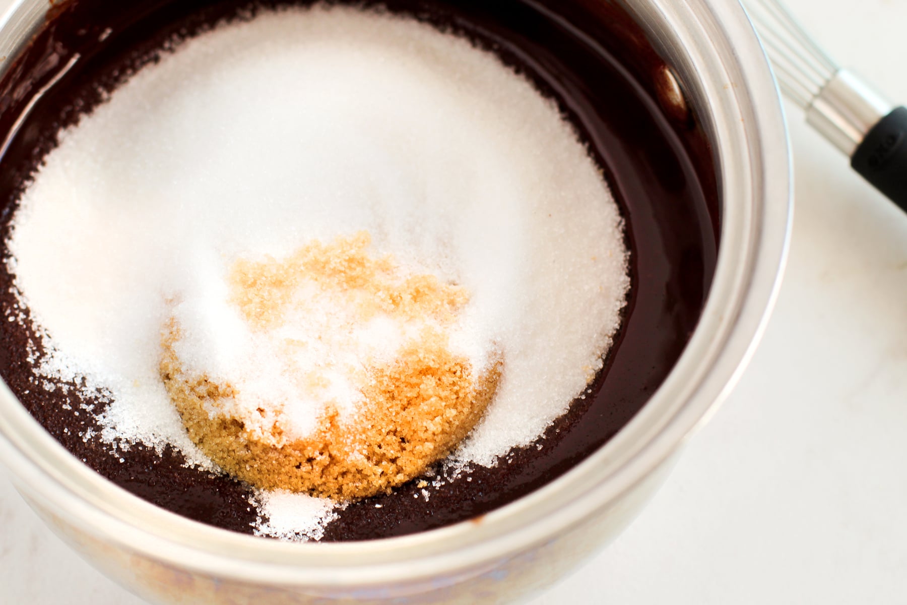 A saucepan containing melted chocolate with piled white and brown sugar on top, and a whisk resting on the side. This is for Fudgy Brownies recipe