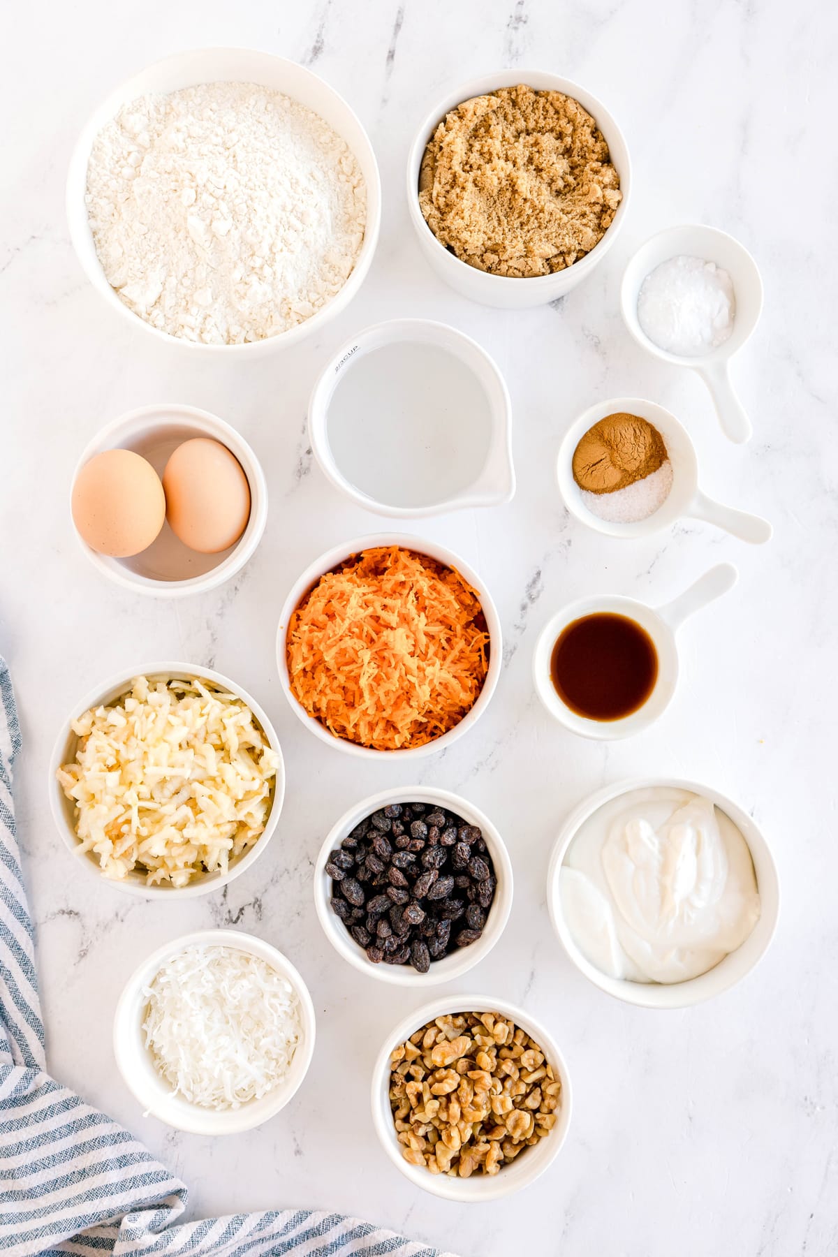 Various baking ingredients in bowls for Morning Glory Muffins, include all purpose flour, brown sugar, baking soda, cinnamon, salt, eggs, coconut oil, sour cream, vanilla extract, grated carrot, grated granny Smith apple, Raisins, Shredded coconut, and chopped walnuts