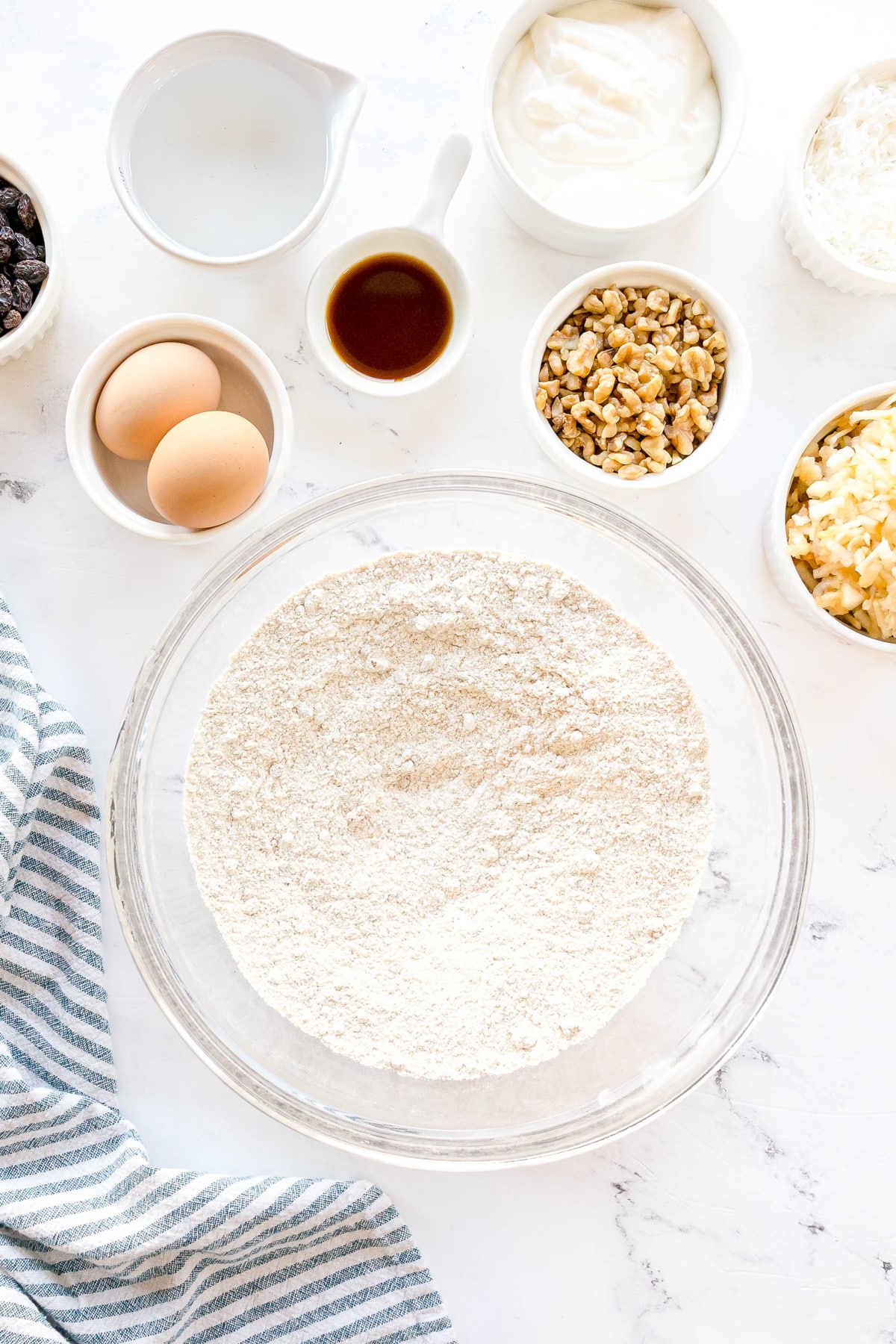 First step in preparing Morning Glory Muffins is to mix flour, brown sugar, baking soda, cinnamon, and salt in a bowl. It is surrounded by small bowls of eggs, walnuts, shredded apples, raisins, yogurt, shredded coconut, vanilla extract, and a cup of water on a white surface.
