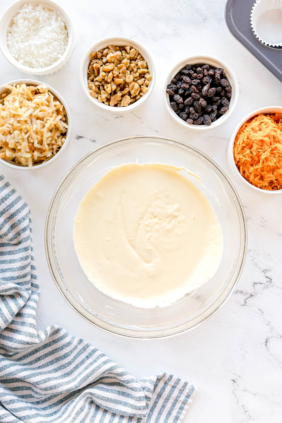 Whisking eggs, coconut oil, sour cream, and vanilla extract until smooth is another step in preparing Morning Glory Muffins. Linen napkins and small bowls of raisins, walnuts and other ingredients can be seen.