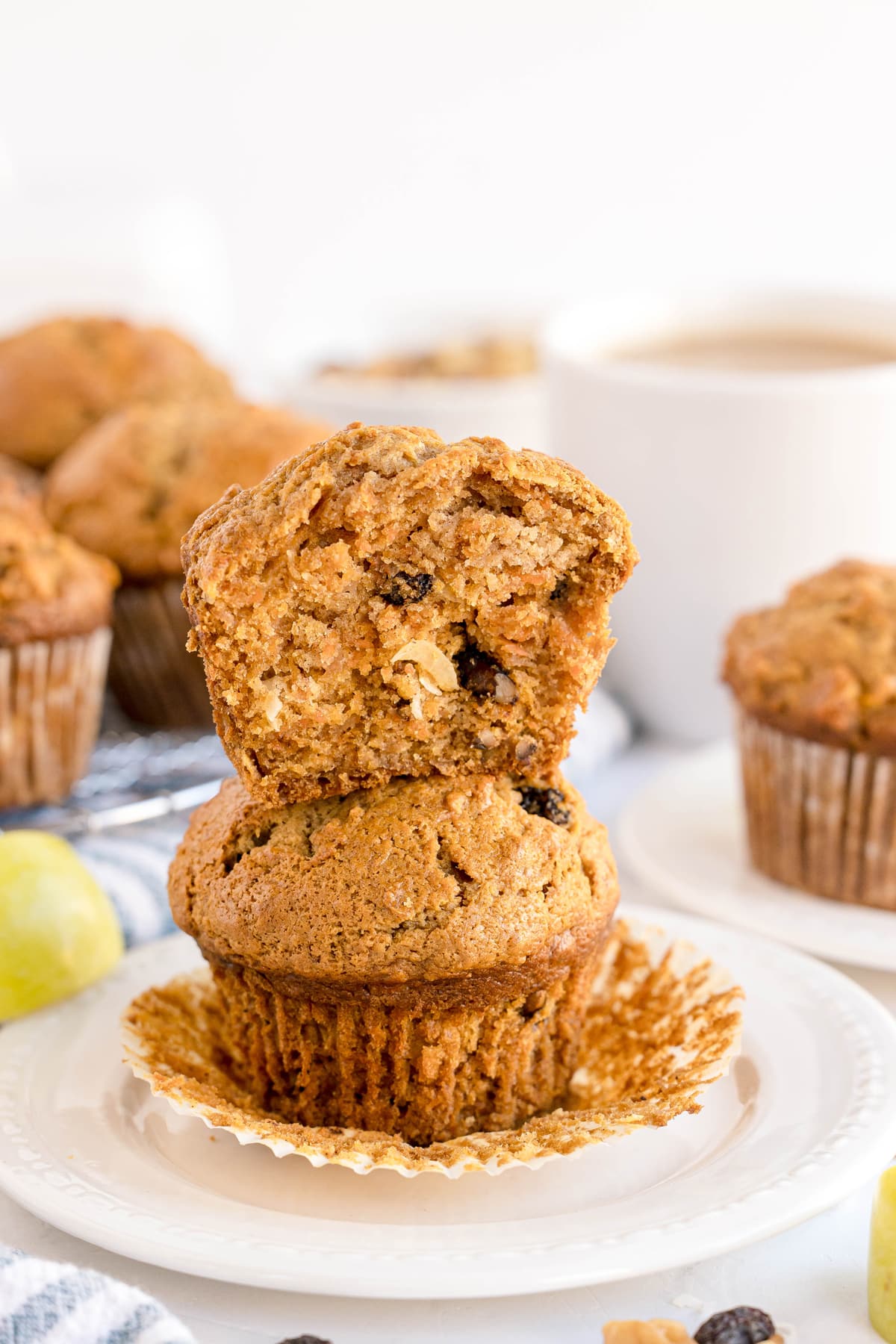 Two Morning Glory Muffins stacked on a white plate, with the top muffin broken open to show its moist, textured interior. Other muffins and a cup of coffee are visible in the background.