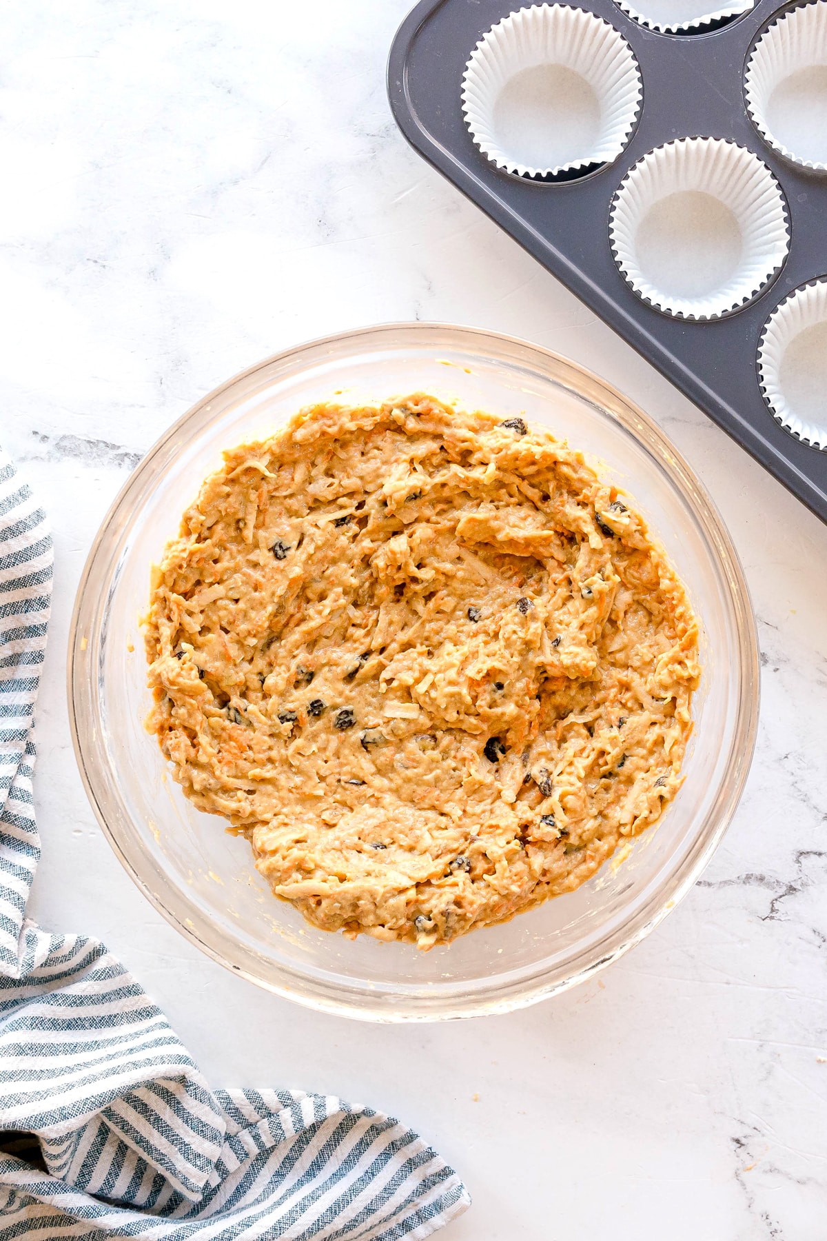 A glass bowl contains thick, mixed Morning Glory Muffins batter with visible raisins. Beside it is a muffin tin lined with white paper cups, set on a white marble surface.