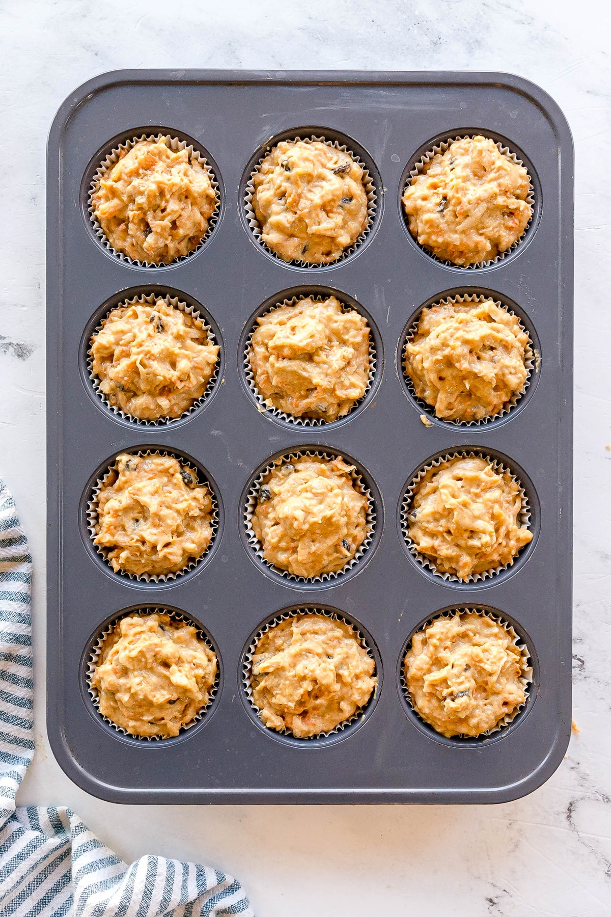 A Morning Glory Muffins tin filled with twelve paper-lined cups, each containing unbaked muffin batter, on a light countertop.