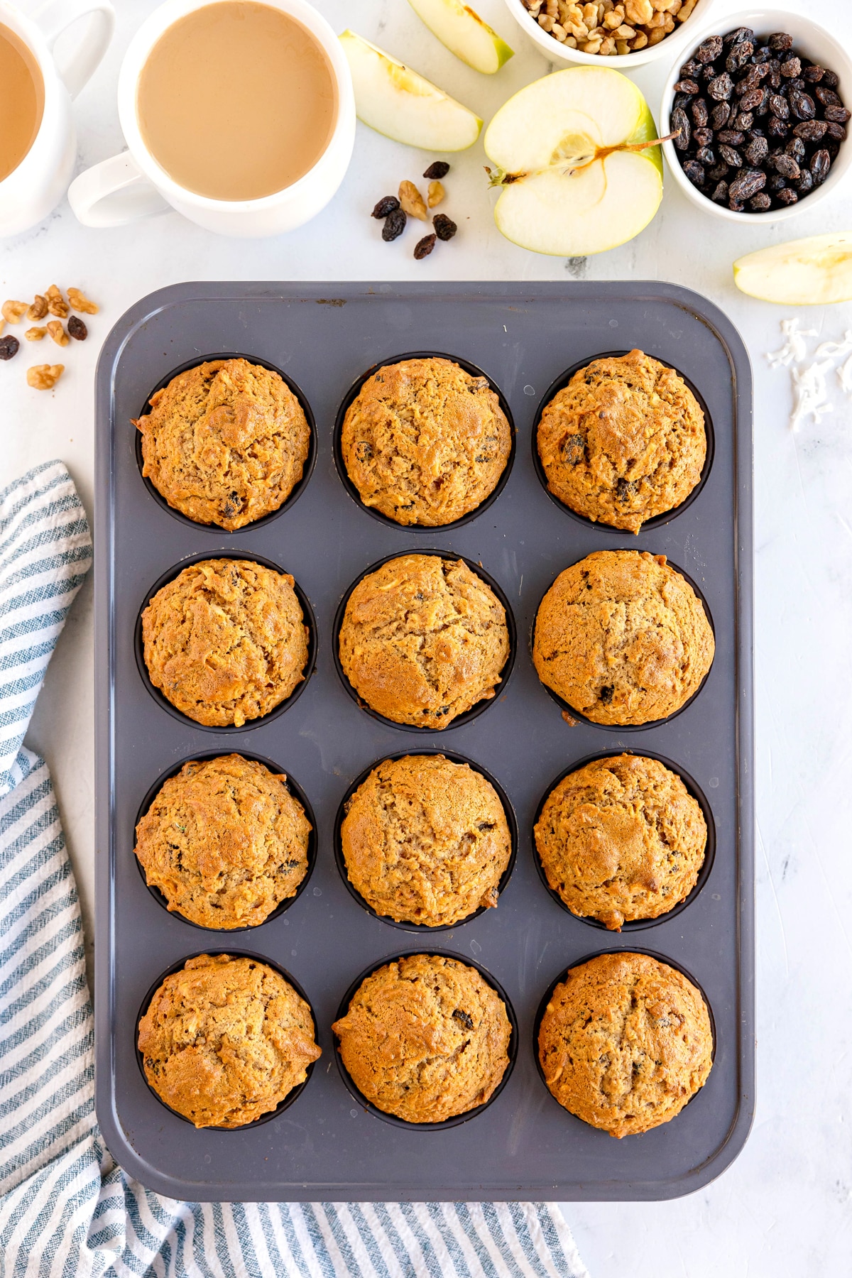 A muffin tin with 12 baked Morning Glory Muffins sits on a counter next to two cups of coffee, apple slices, walnuts, raisins, and a blue striped towel.