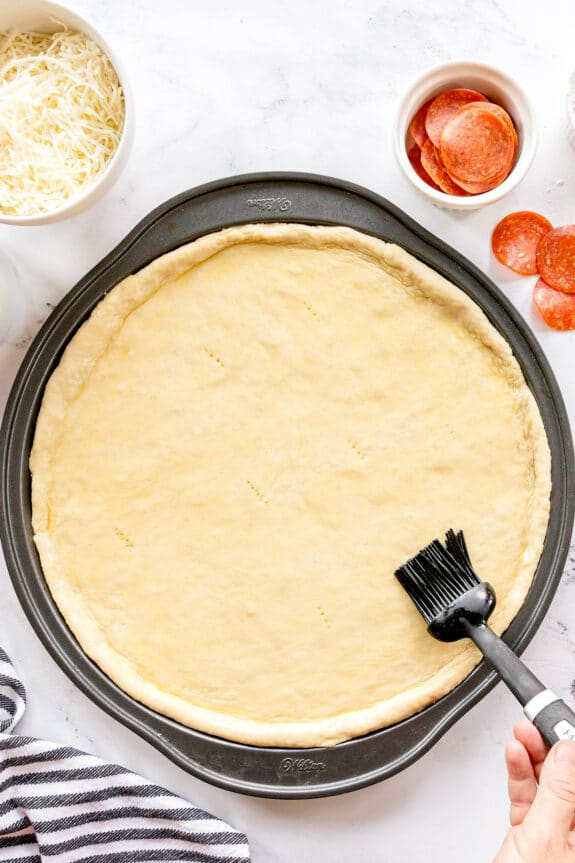 A hand brushes oil or butter onto a round pizza dough in a pan, with bowls of shredded cheese and pepperoni nearby on a white surface—perfect prep for homemade pepperoni pizza.