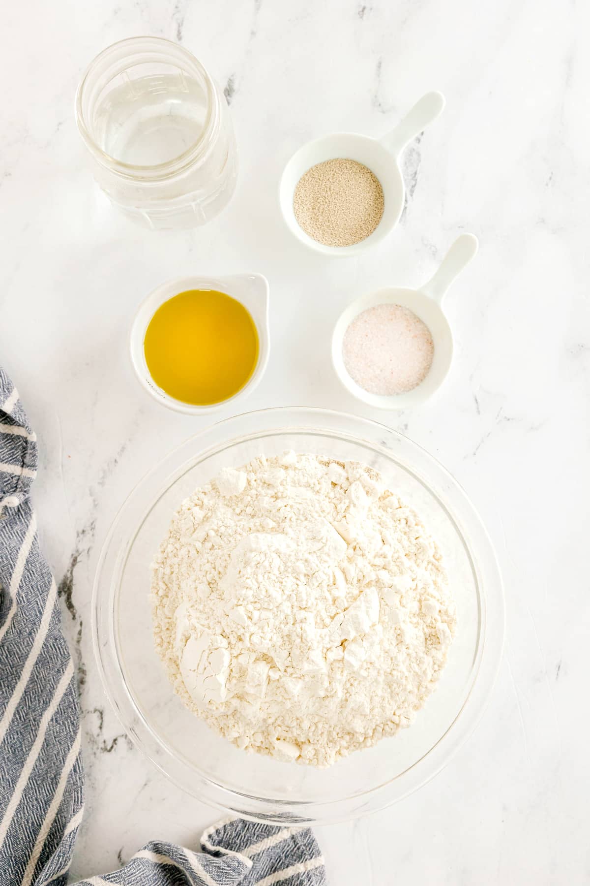 Ingredient of Pizza Dough on a white counter include: a glass bowl of flour, a small jar of water, a bowl of virgin olive oil, a bowl of active dry yeast, and a bowl of kosher salt on a marble surface with a striped towel nearby.