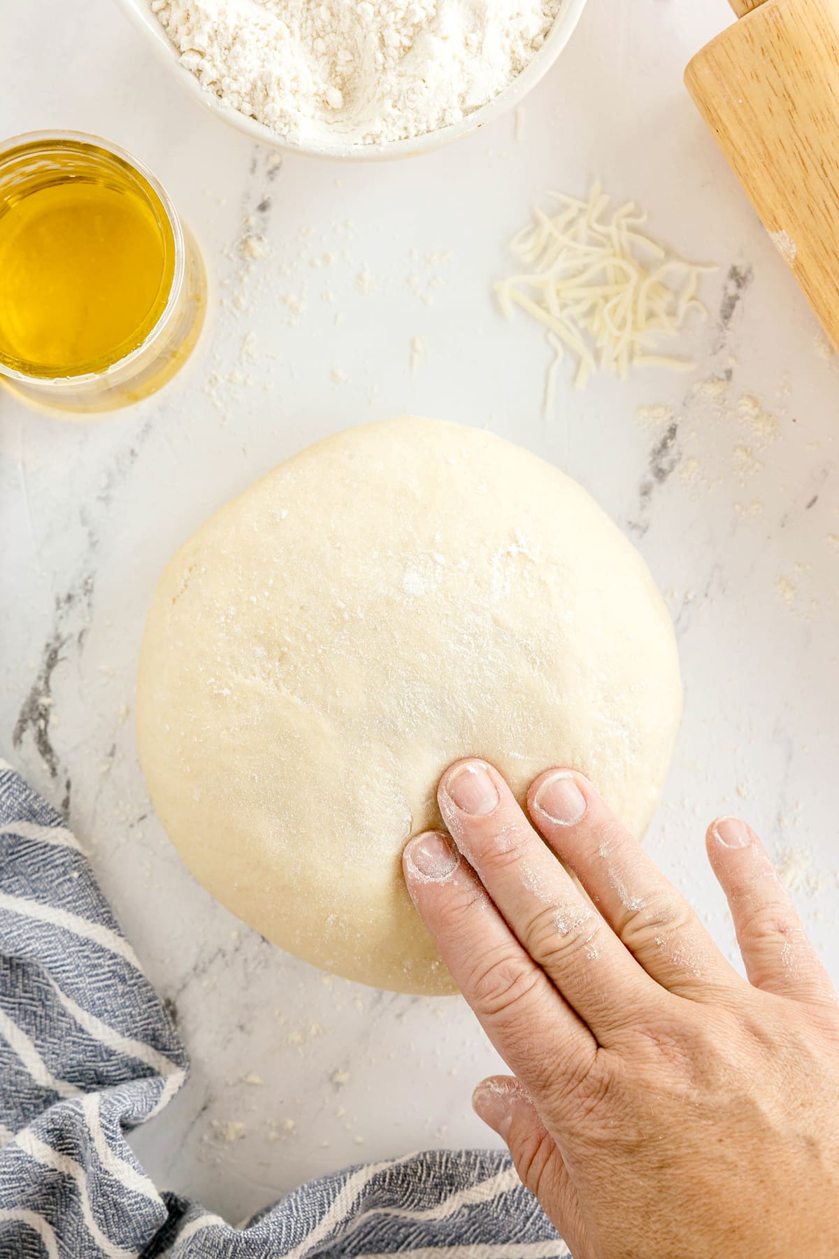 A hand presses on a ball of pizza dough on a floured surface, with a jar of oil, a bowl of flour, and shredded cheese nearby.