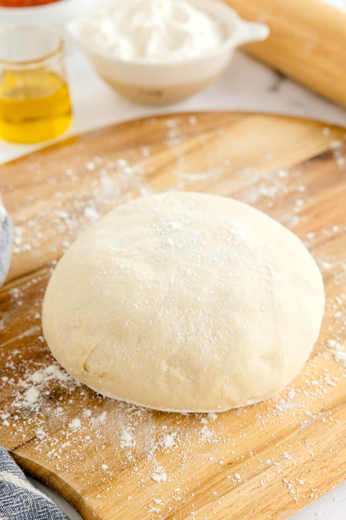 A round ball of raw pizza dough dusted with flour rests on a wooden board, with olive oil, flour, and a rolling pin in the background.