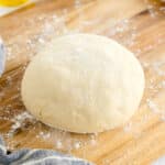 A round ball of raw Pizza dough dusted with flour sits on a floured wooden surface next to a striped cloth.